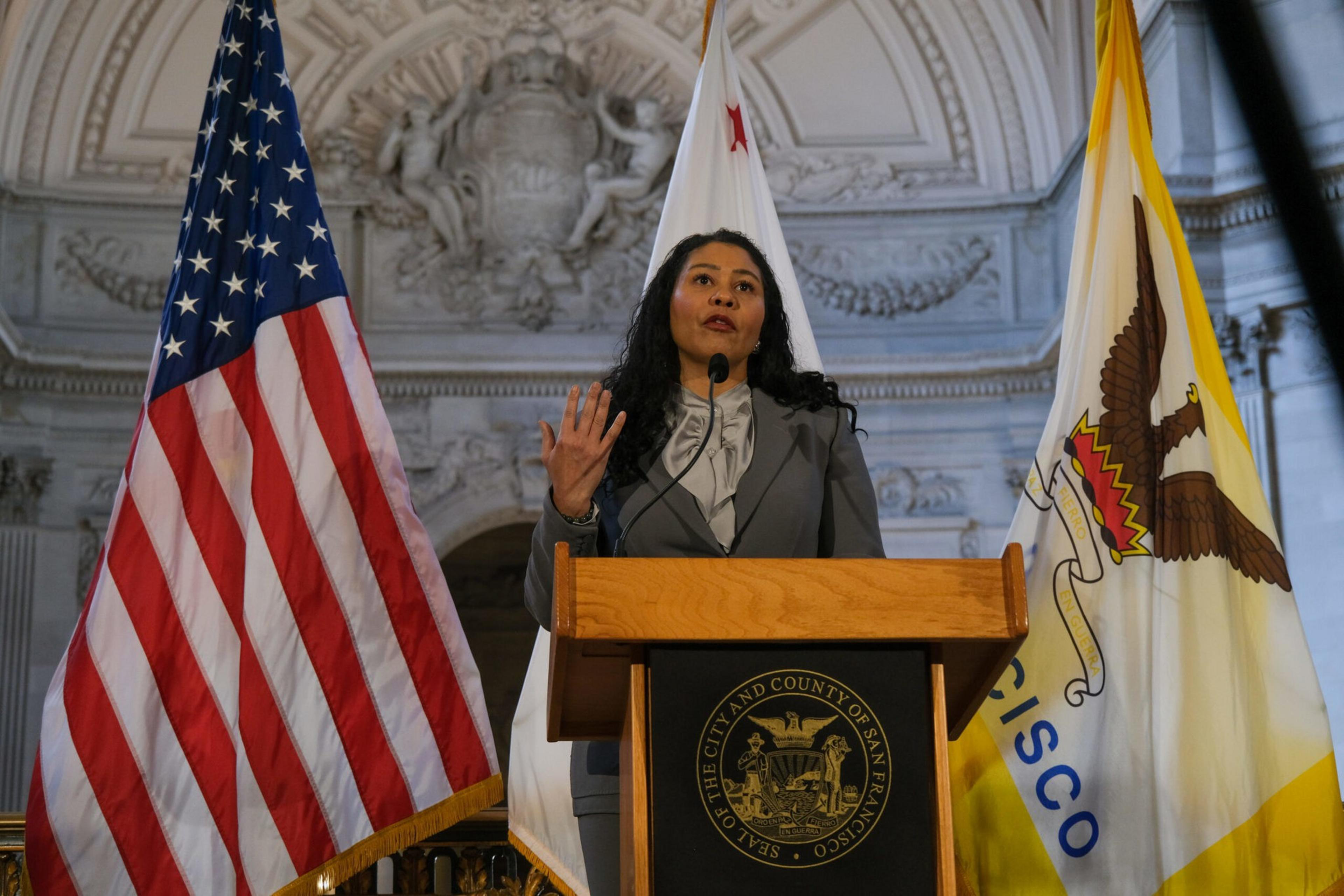 A woman in a gray suit stands behind a podium with the San Francisco seal, flanked by American and San Francisco flags, speaking in a grand hall.