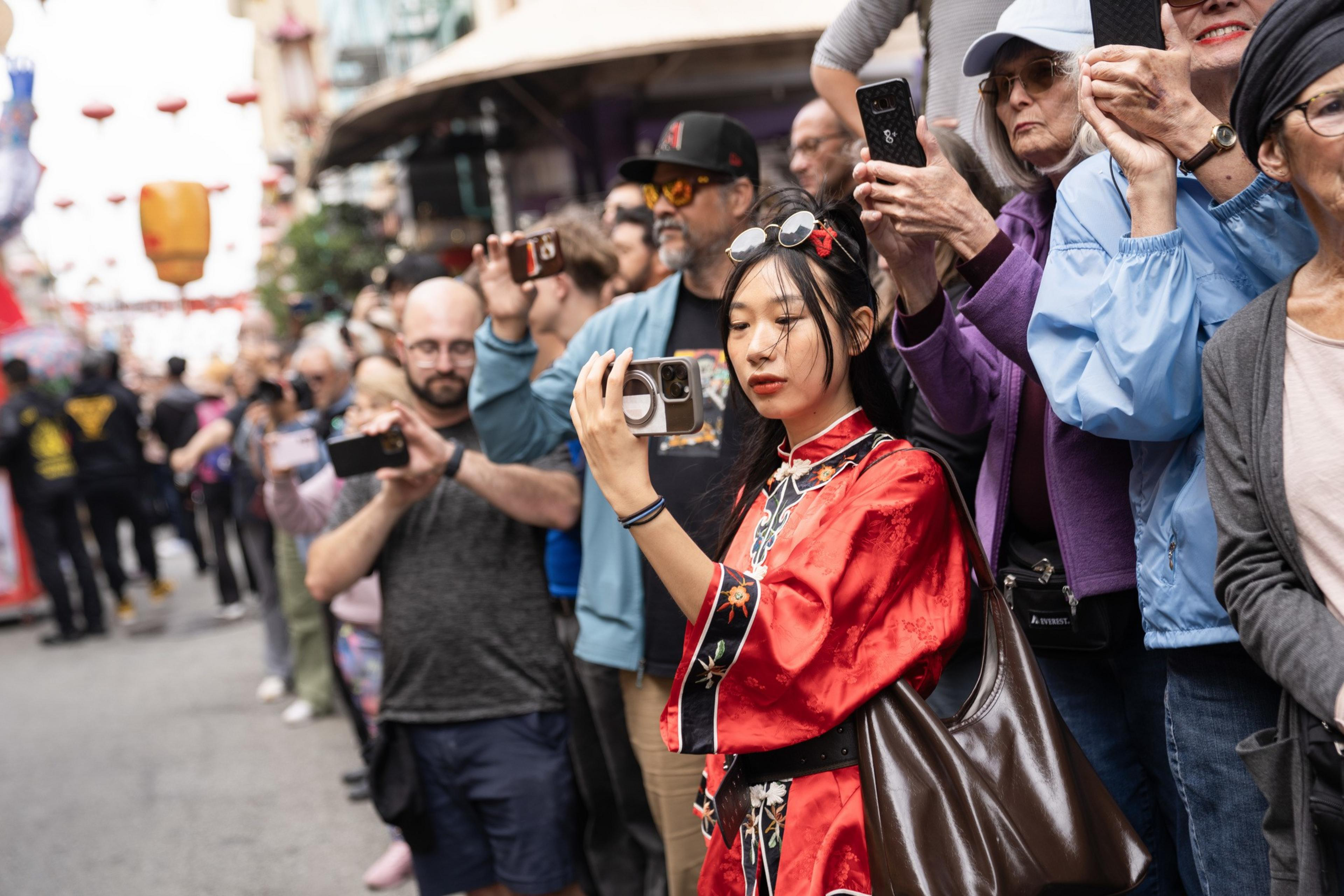 A diverse crowd stands closely together on a street, many holding phones and cameras, with a woman in a red embroidered outfit prominently capturing a moment.