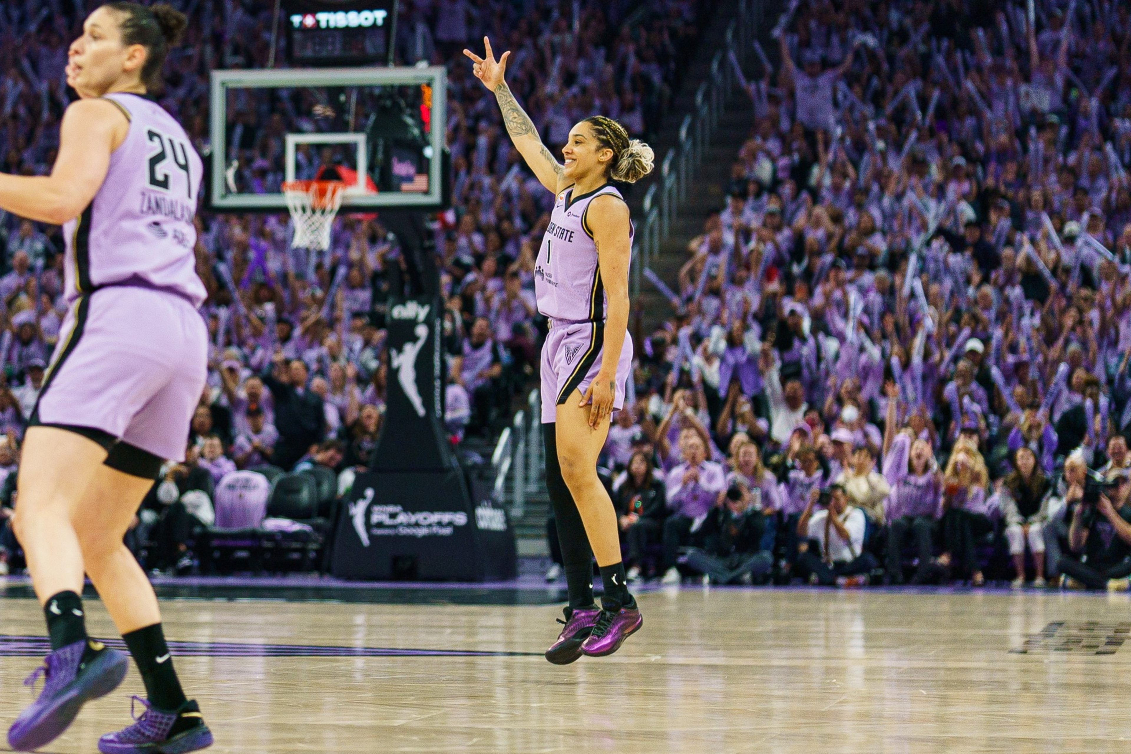 A female basketball player in a purple uniform jumps with one arm raised, celebrating as a crowd cheers wildly in the background.