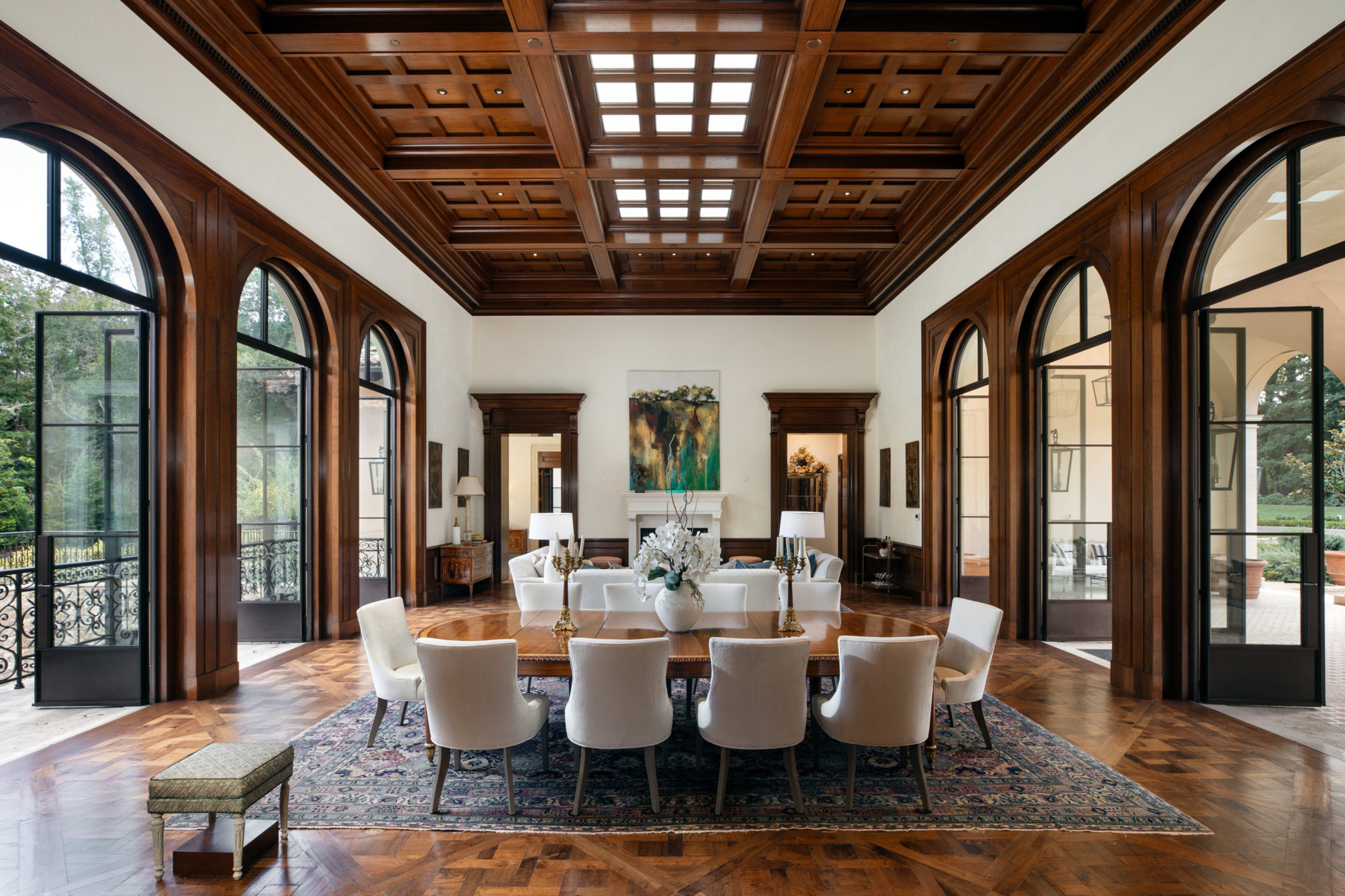 A spacious dining room features a large wooden table with eight cream chairs, a floral centerpiece, wooden coffered ceiling, arched windows, and hardwood floors.