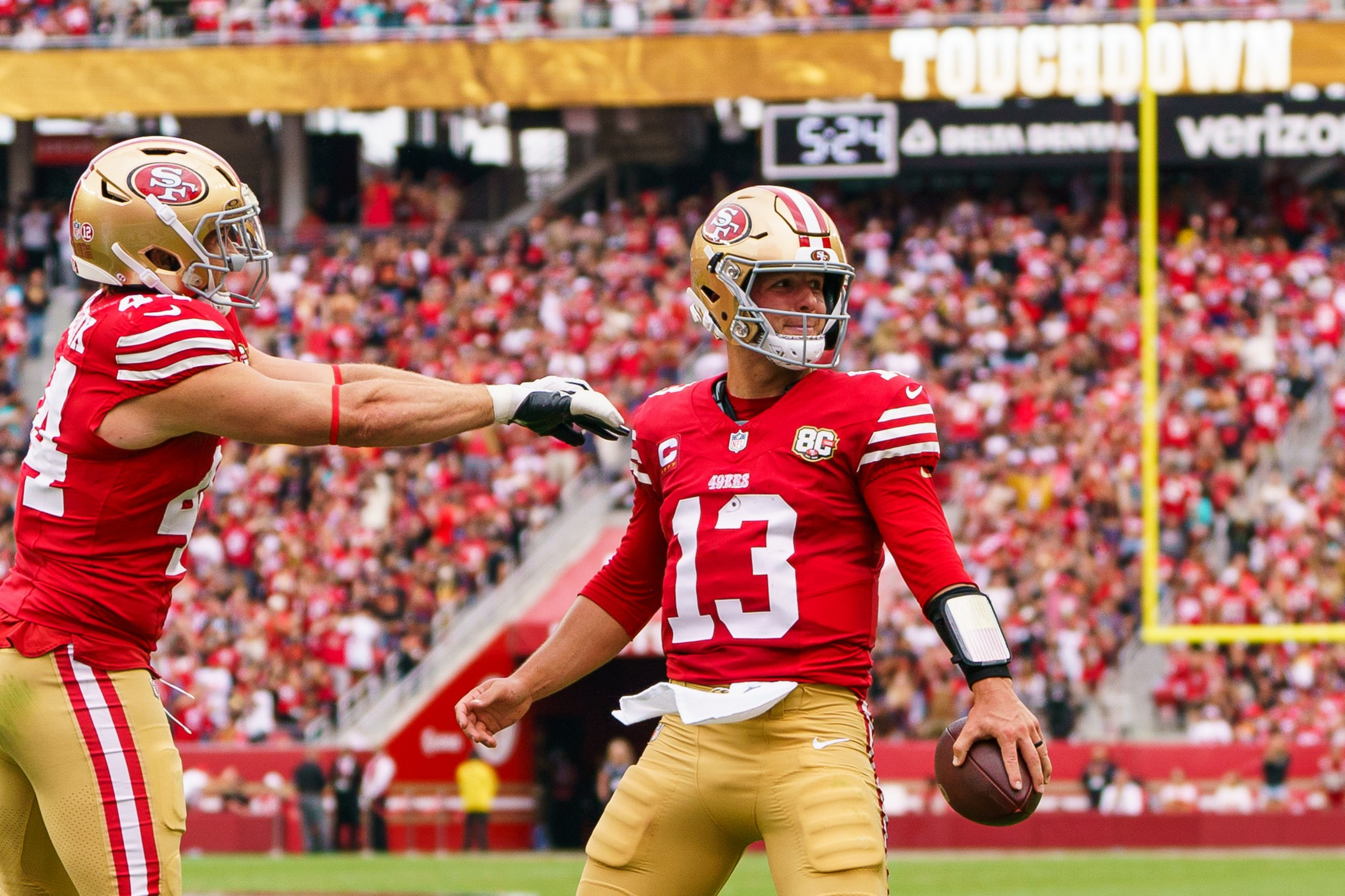 A San Francisco 49ers quarterback wearing number 13 prepares to throw a football while a teammate reaches out to him on the field.