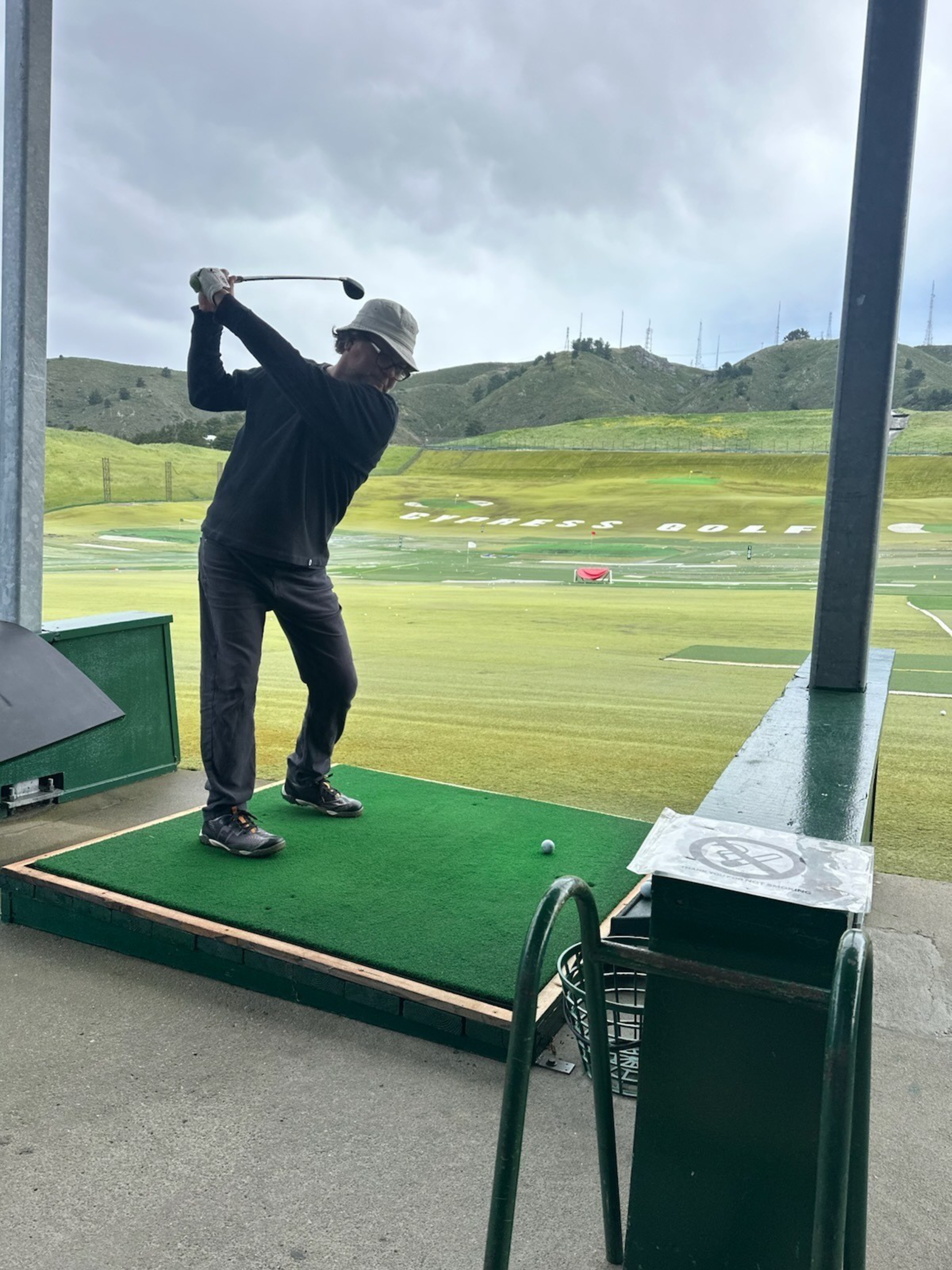 A person in a hat and dark clothing is mid-swing at a golf driving range, standing on a green mat with a golf ball in front of them.