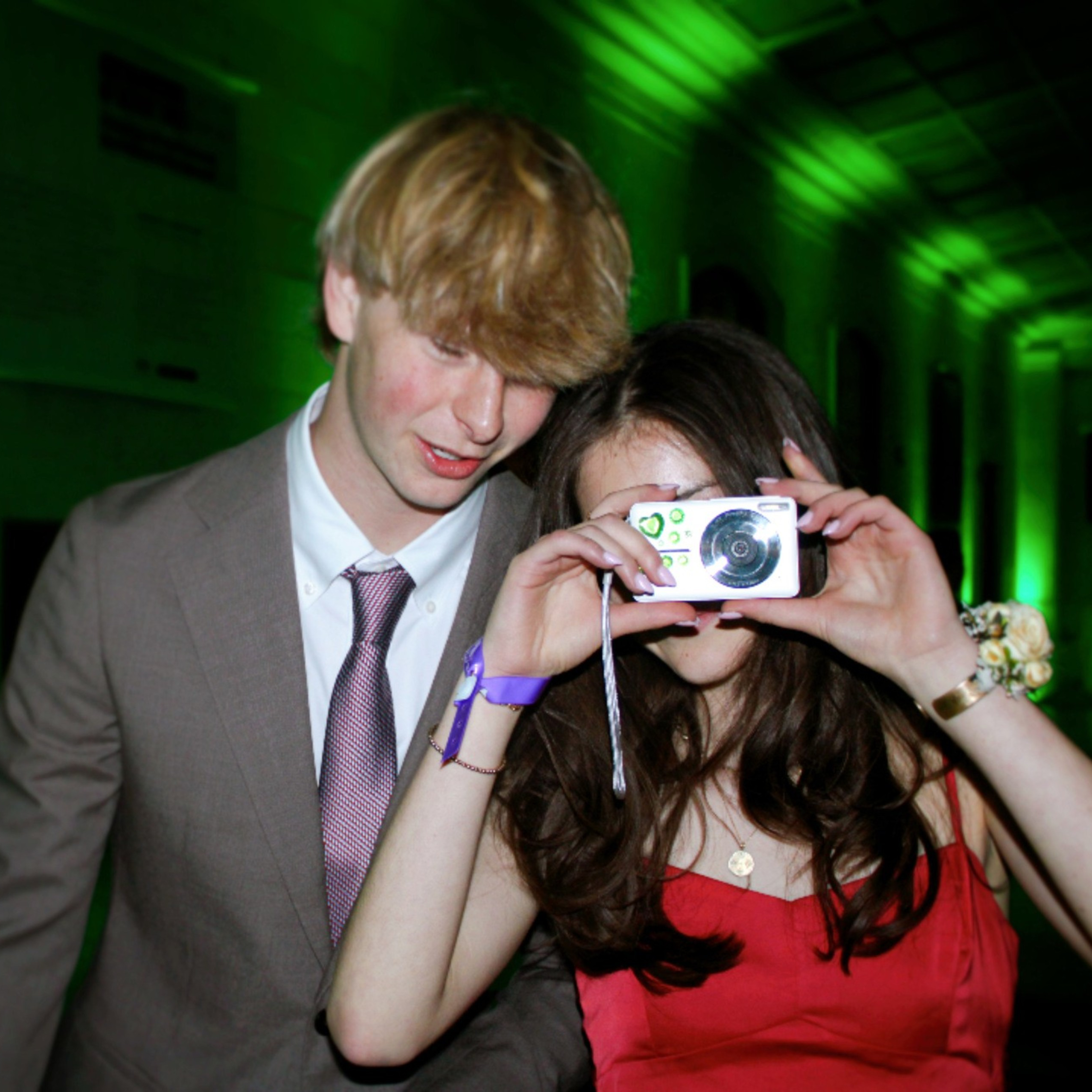 A young woman in a red dress holds a camera to her face, standing beside a young man in a suit and tie. The background is illuminated with green lighting.