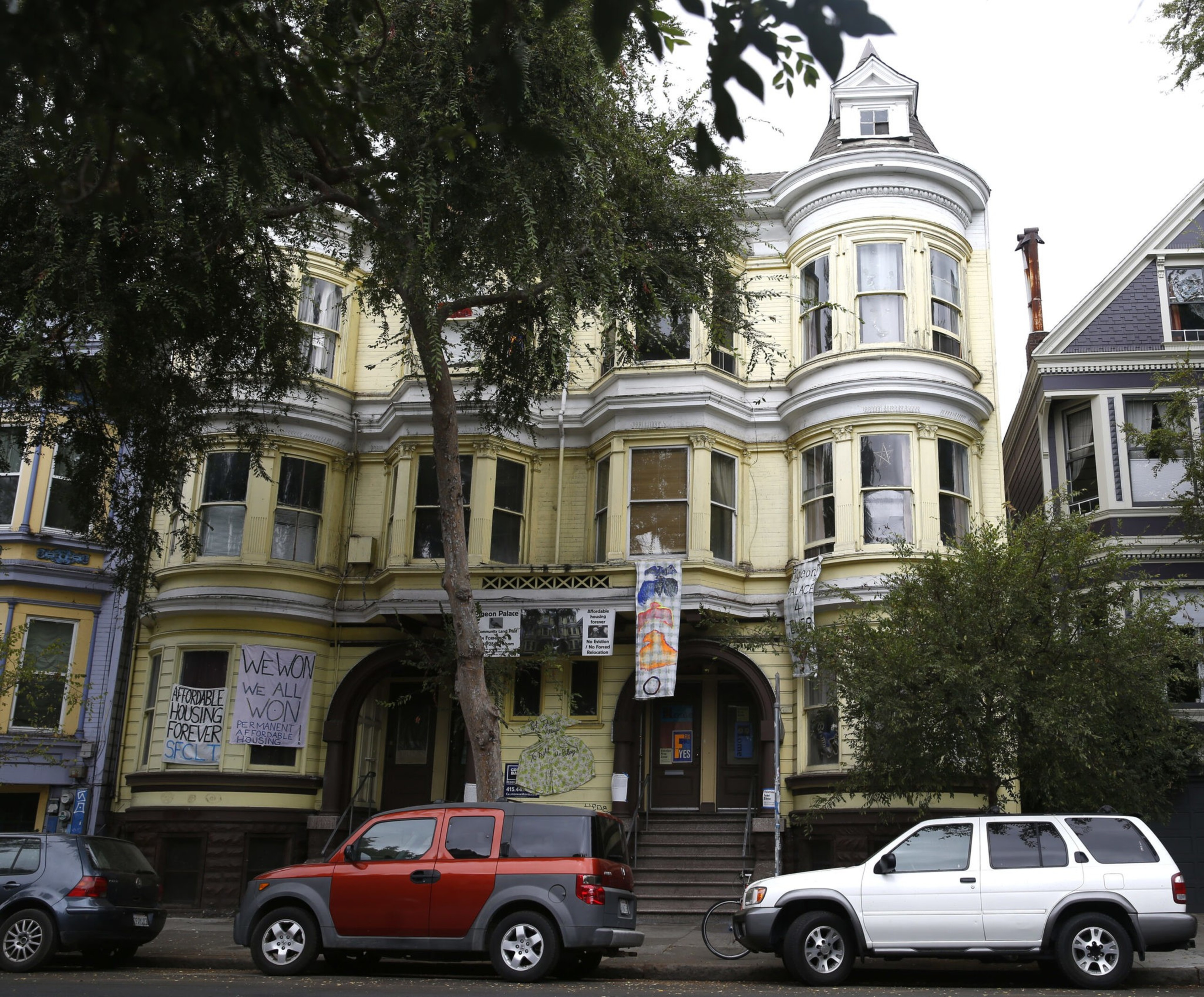A yellow Victorian-style building with banners advocating affordable housing hangs above its entrance, while three cars are parked on the street in front.
