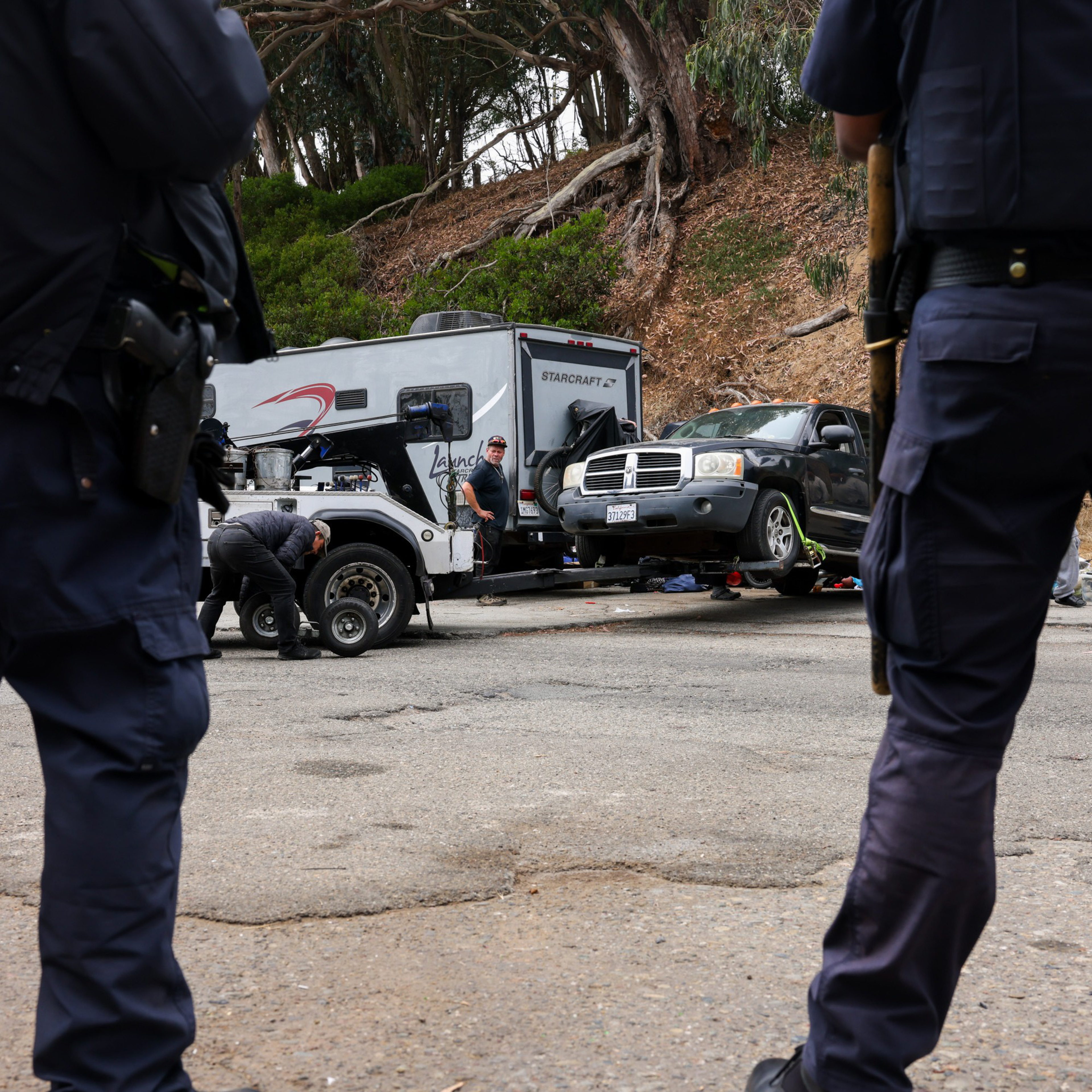 Several police officers stand near a vehicle being serviced beside a trailer and a pickup truck on a forested roadside. Tools and equipment are scattered around.