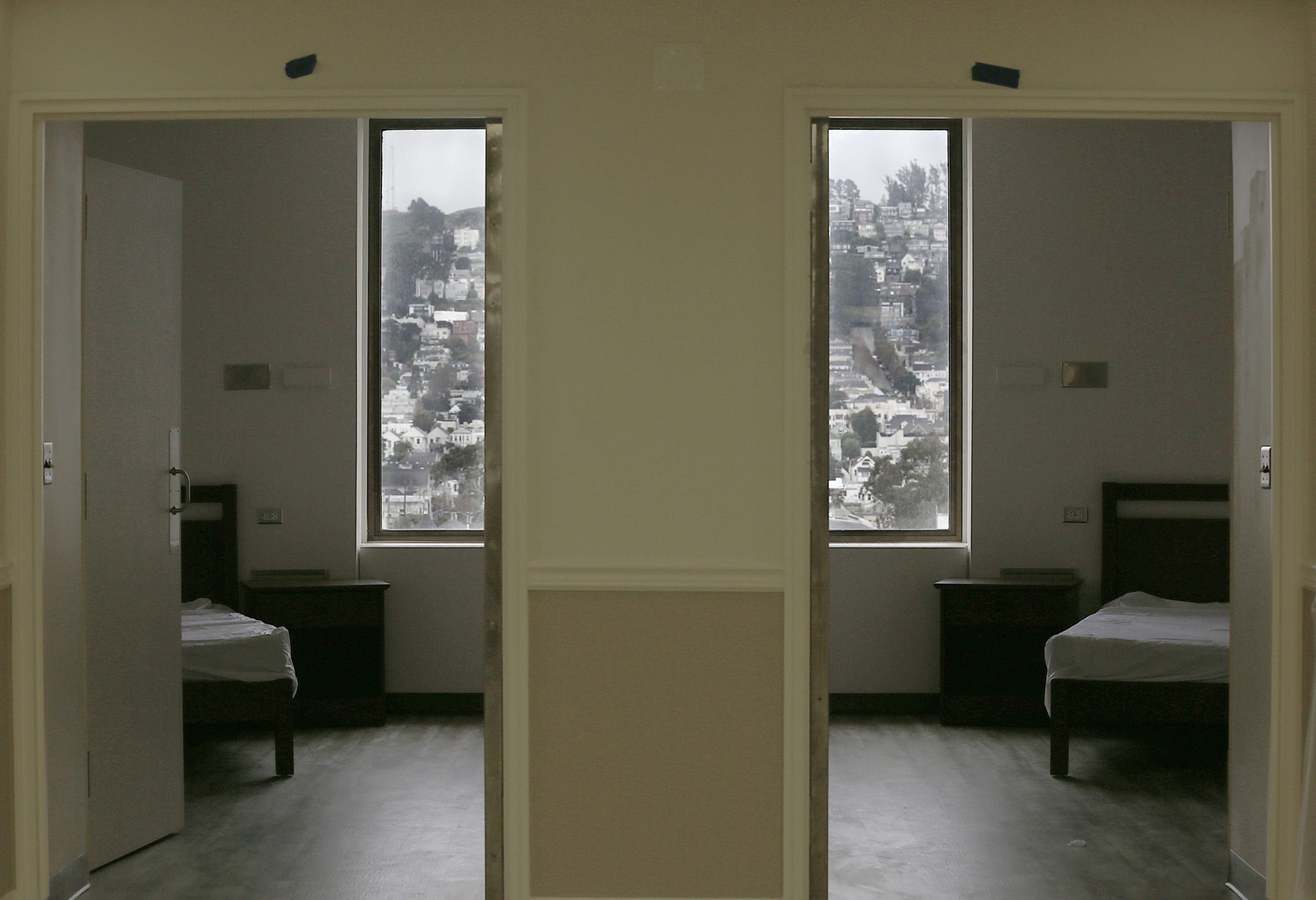 Two identical, sparsely furnished rooms with single beds and nightstands are seen through adjacent open doorways, each featuring a window with a cityscape view.