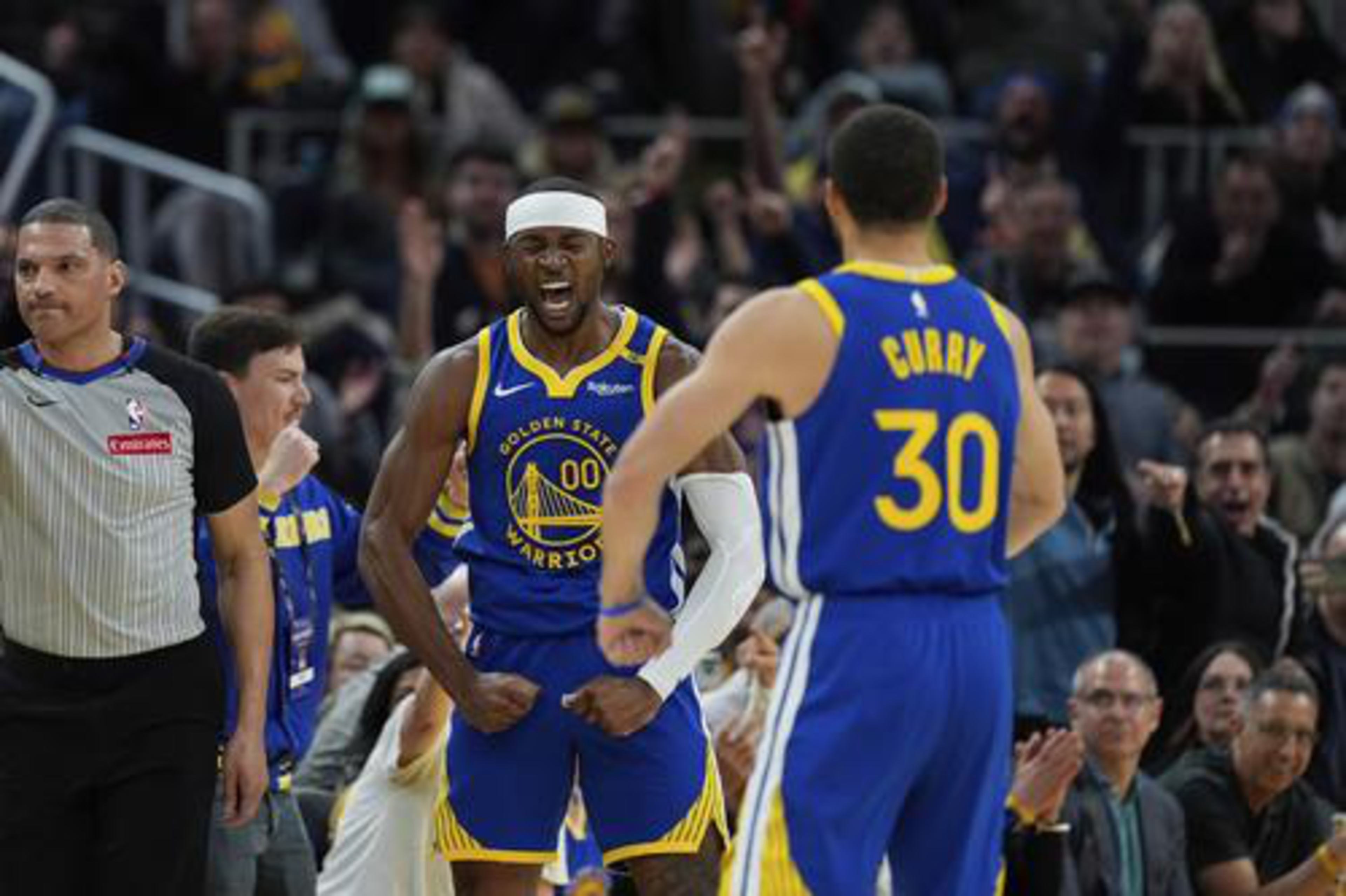 A Golden State Warriors player celebrates intensely while facing teammate Curry, with a referee and cheering crowd in the background.