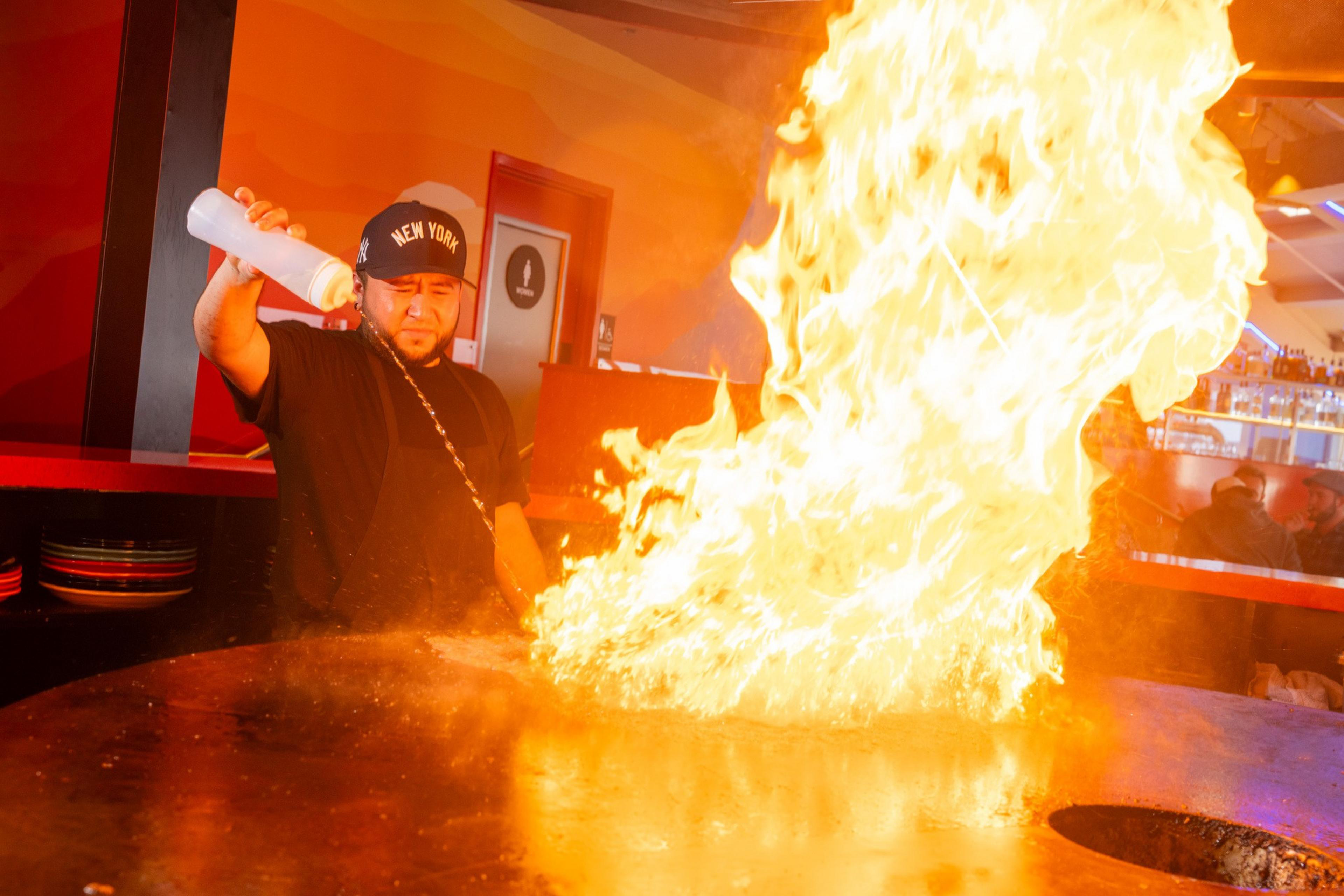 A chef wearing a New York cap creates a large flame burst on a round cooking surface inside a restaurant.