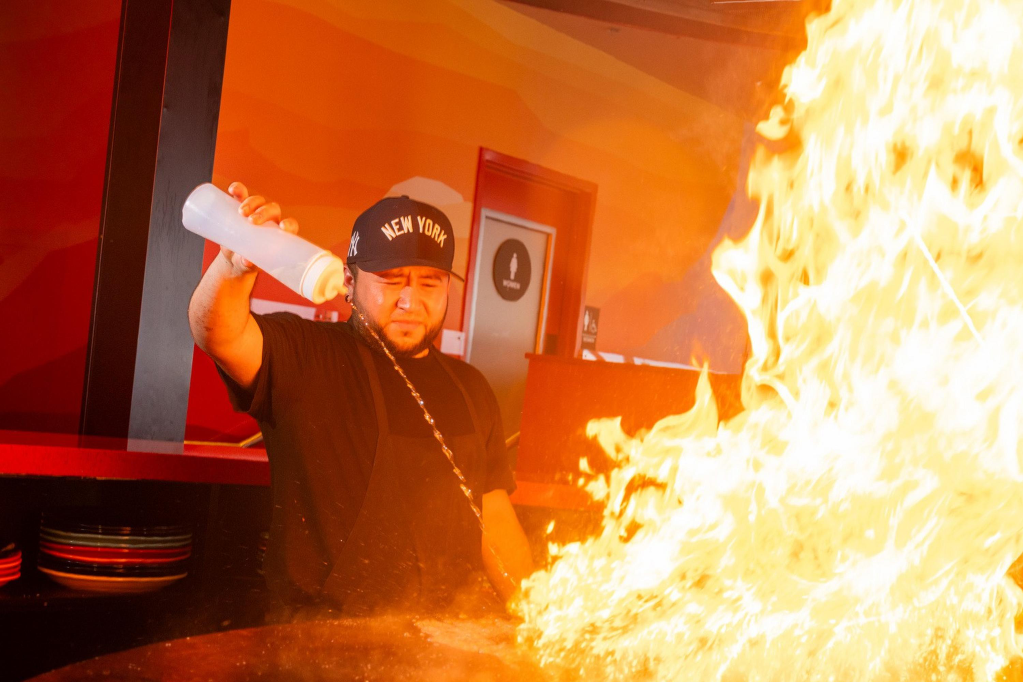 A chef wearing a New York cap creates a large flame burst on a round cooking surface inside a restaurant.