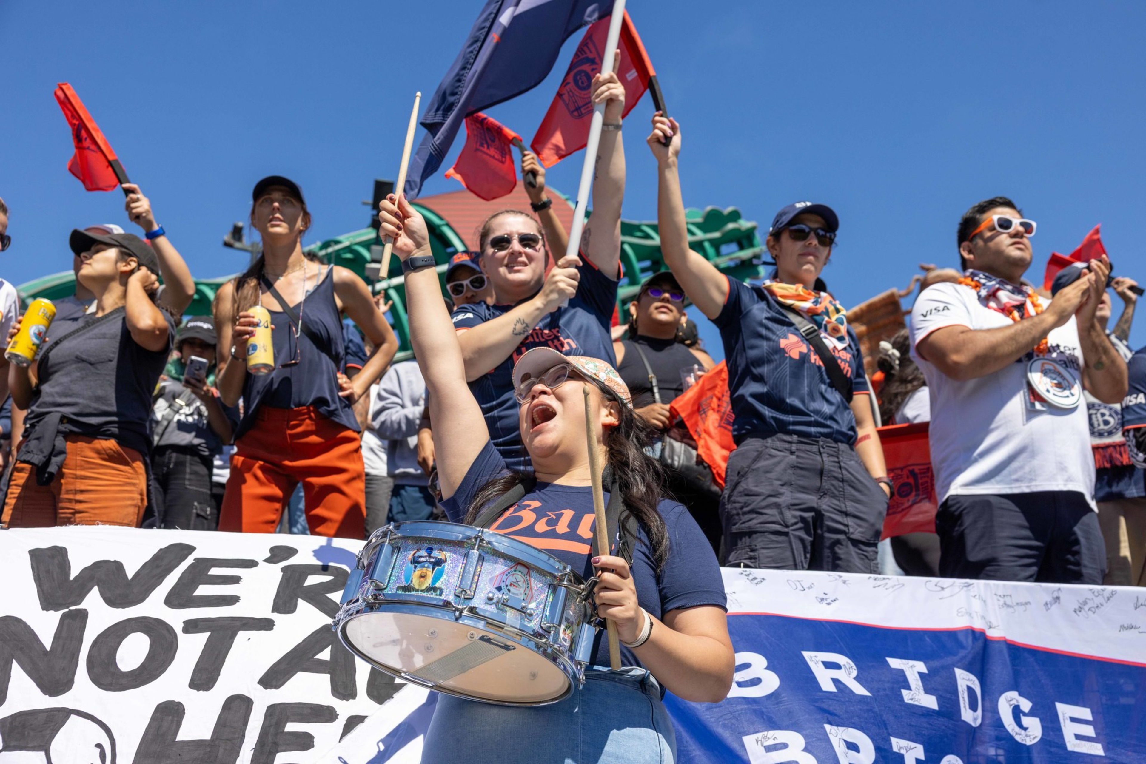 A diverse crowd cheers energetically, waving flags and holding drinks, while a woman in the foreground beats a drum enthusiastically.