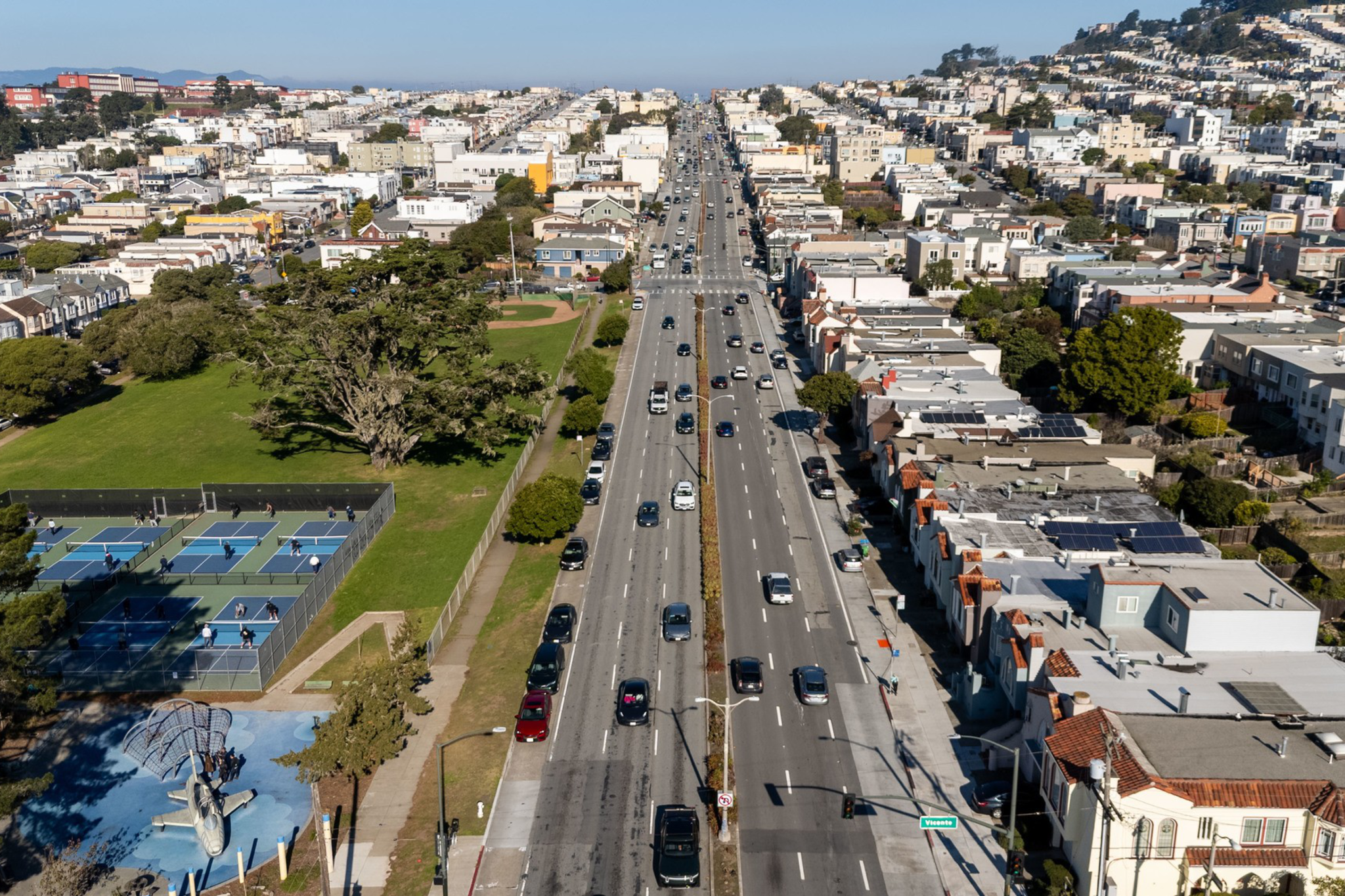 A wide street flanked by residential buildings on one side and a park with tennis courts on the other stretches into the distance under a clear blue sky.
