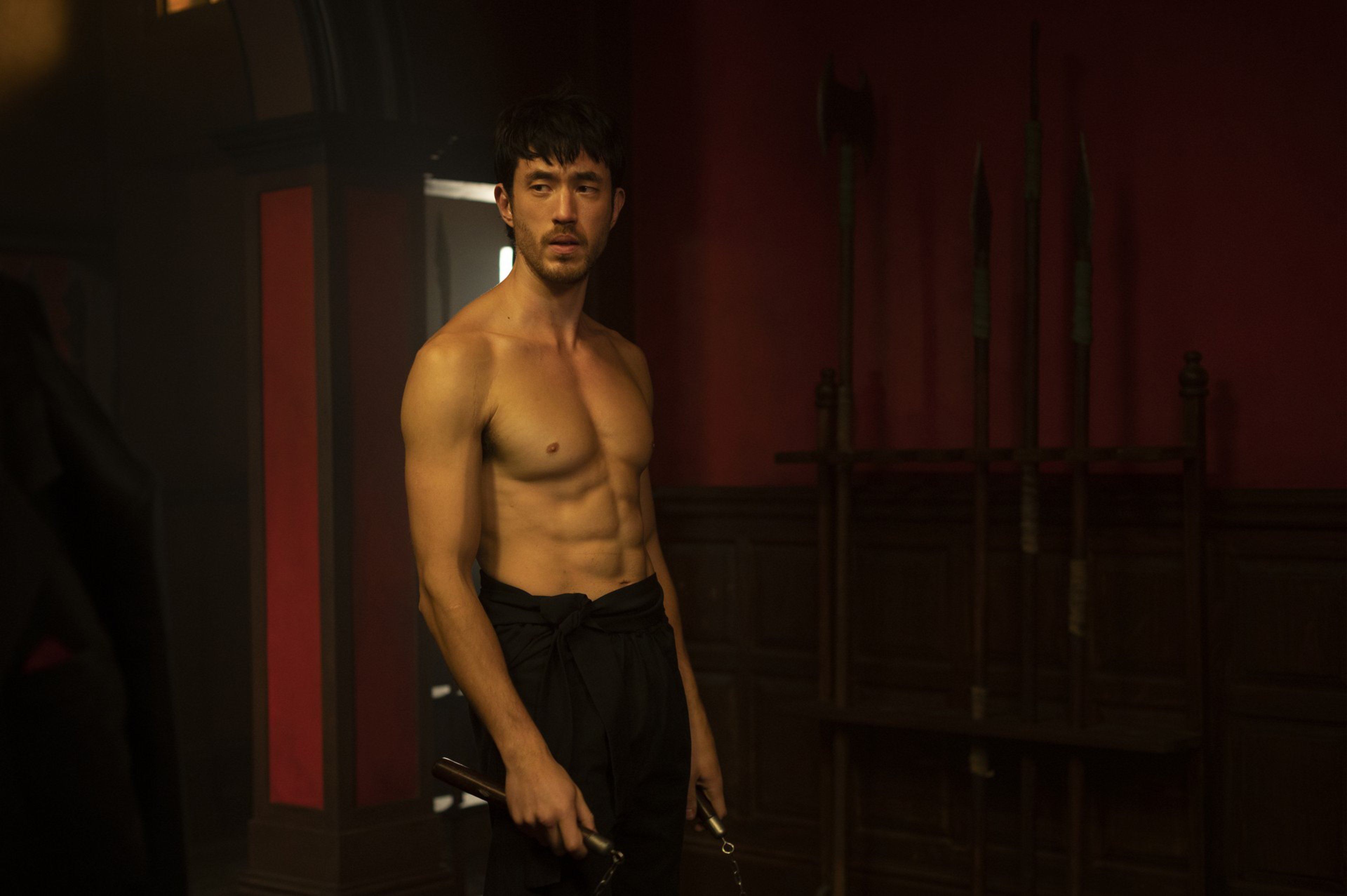A shirtless man with a muscular build stands in a dimly lit room holding nunchaku, with wooden spears displayed on a rack behind him.