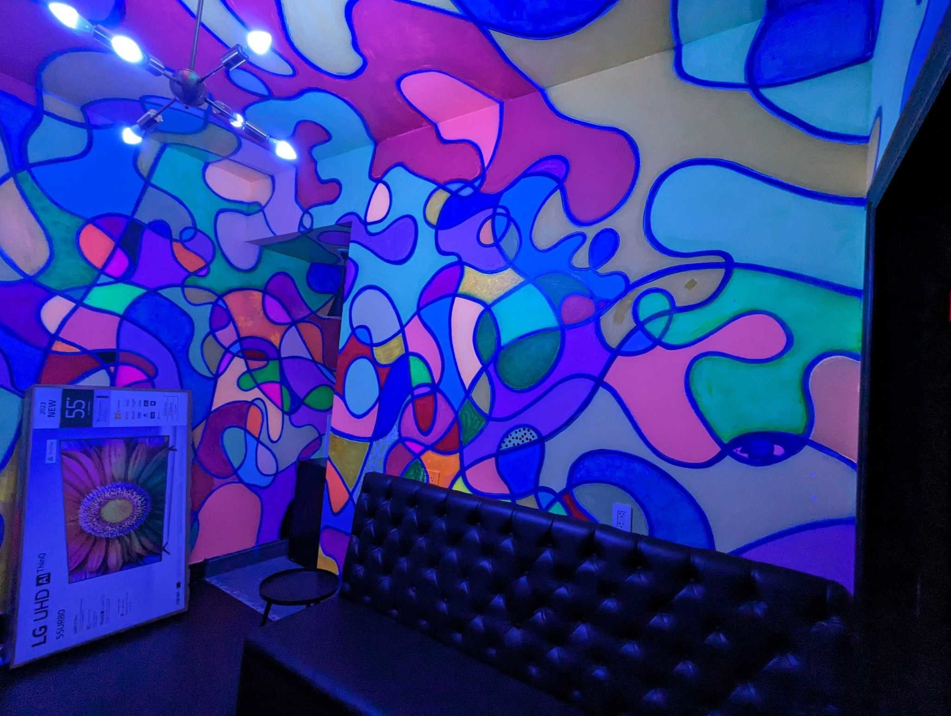 A room with abstract shapes on the wall, a black sofa, a ceiling light, and a TV in a box.