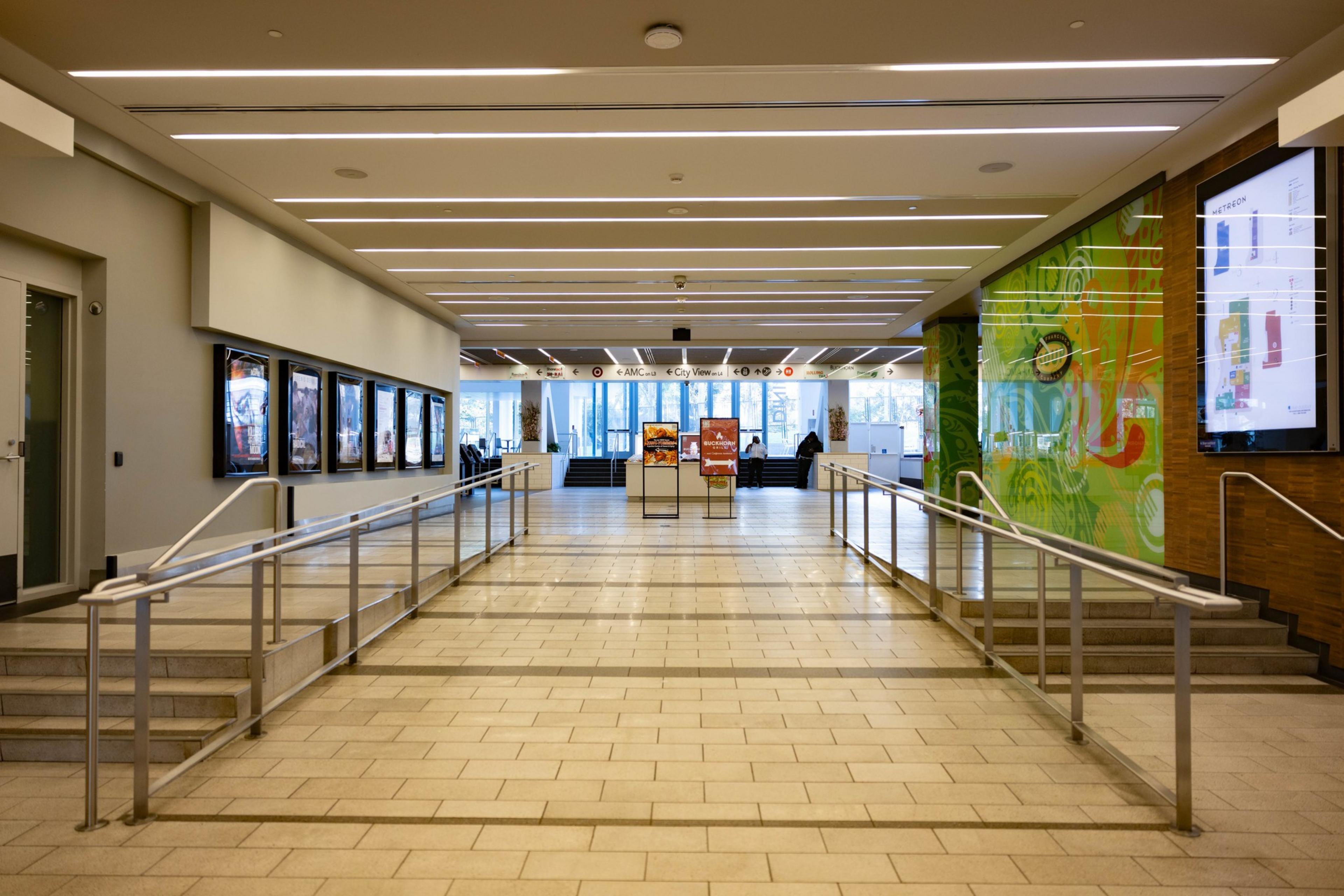 A spacious hallway with a tiled floor, accented by railings on either side, leading to a sunlit area with signs and large windows. Walls display colorful posters.