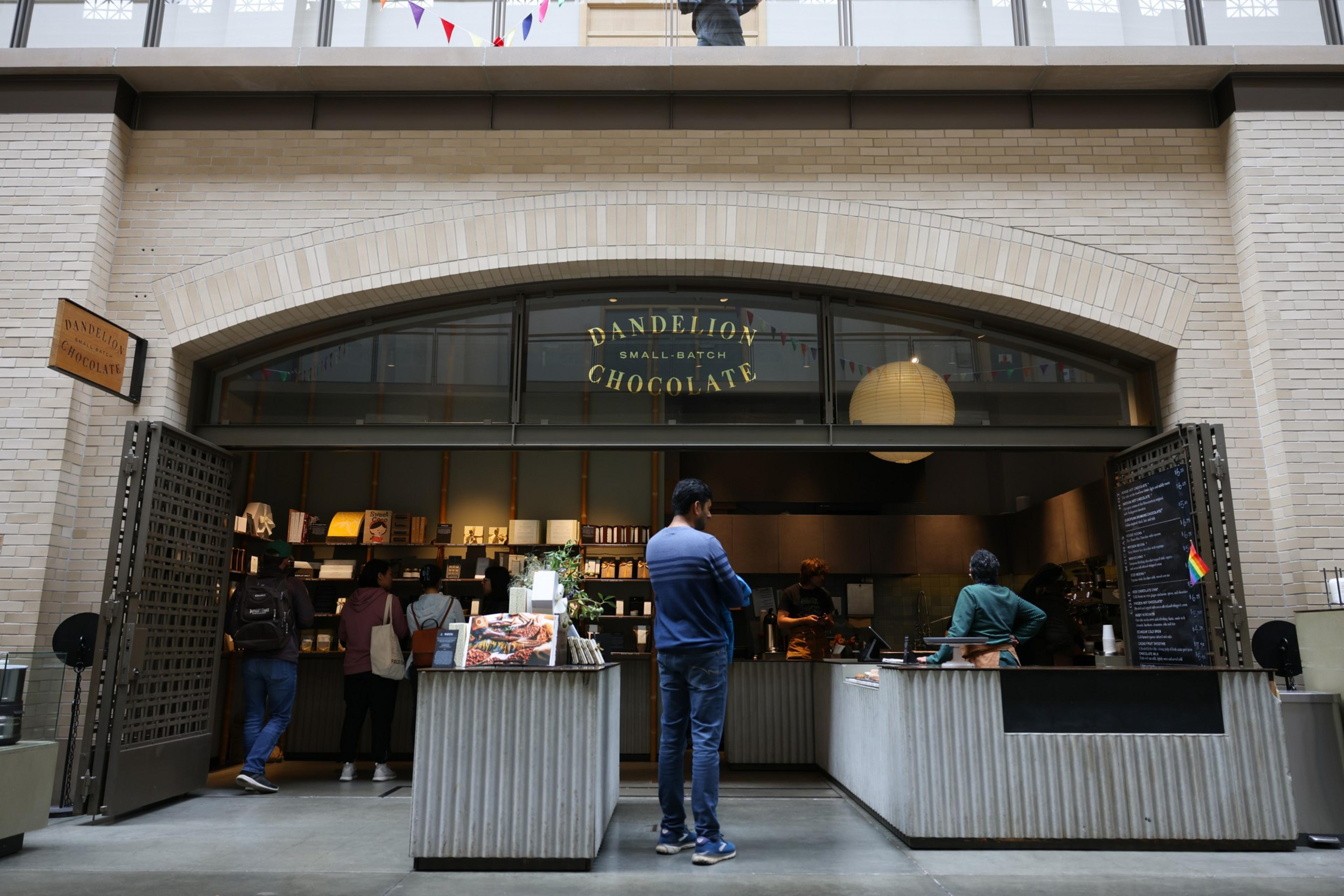A small chocolate shop with a tan brick arch, open metal gates, customers inside, and a man in a blue sweater standing at the counter.
