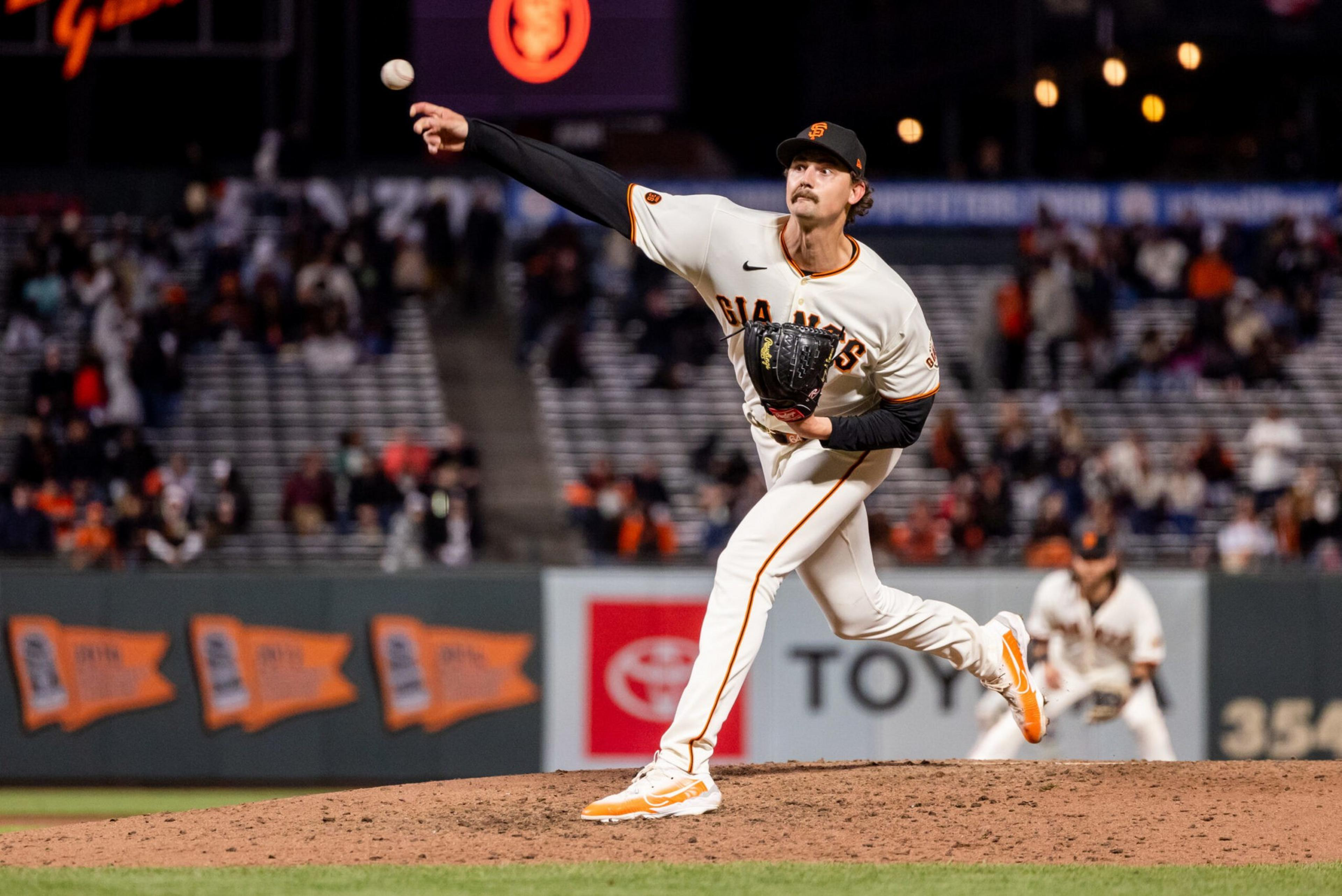 A baseball pitcher in a San Francisco Giants uniform is throwing a pitch during a game, with a teammate ready in the background.