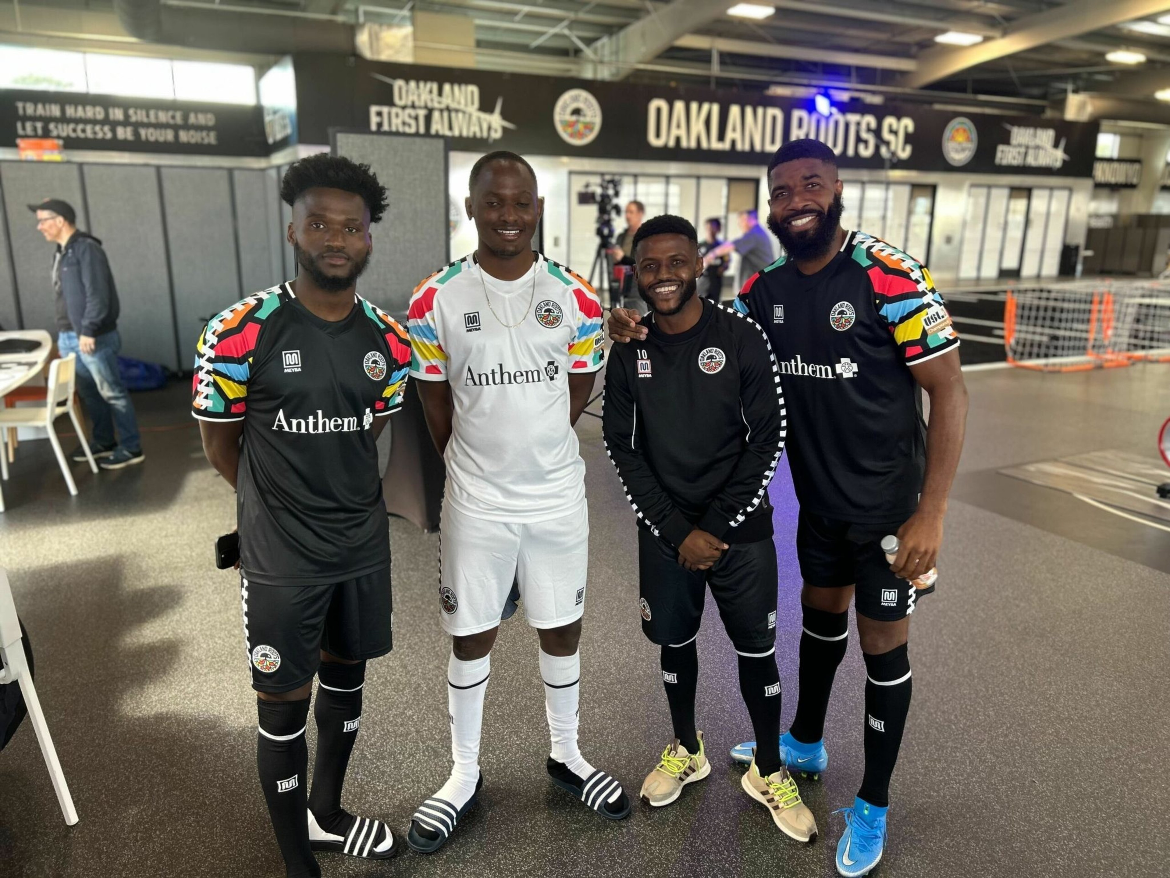 Four men stand indoors wearing Oakland Roots SC soccer uniforms, three in black kits and one in a white kit, smiling for the camera with a sports center background.