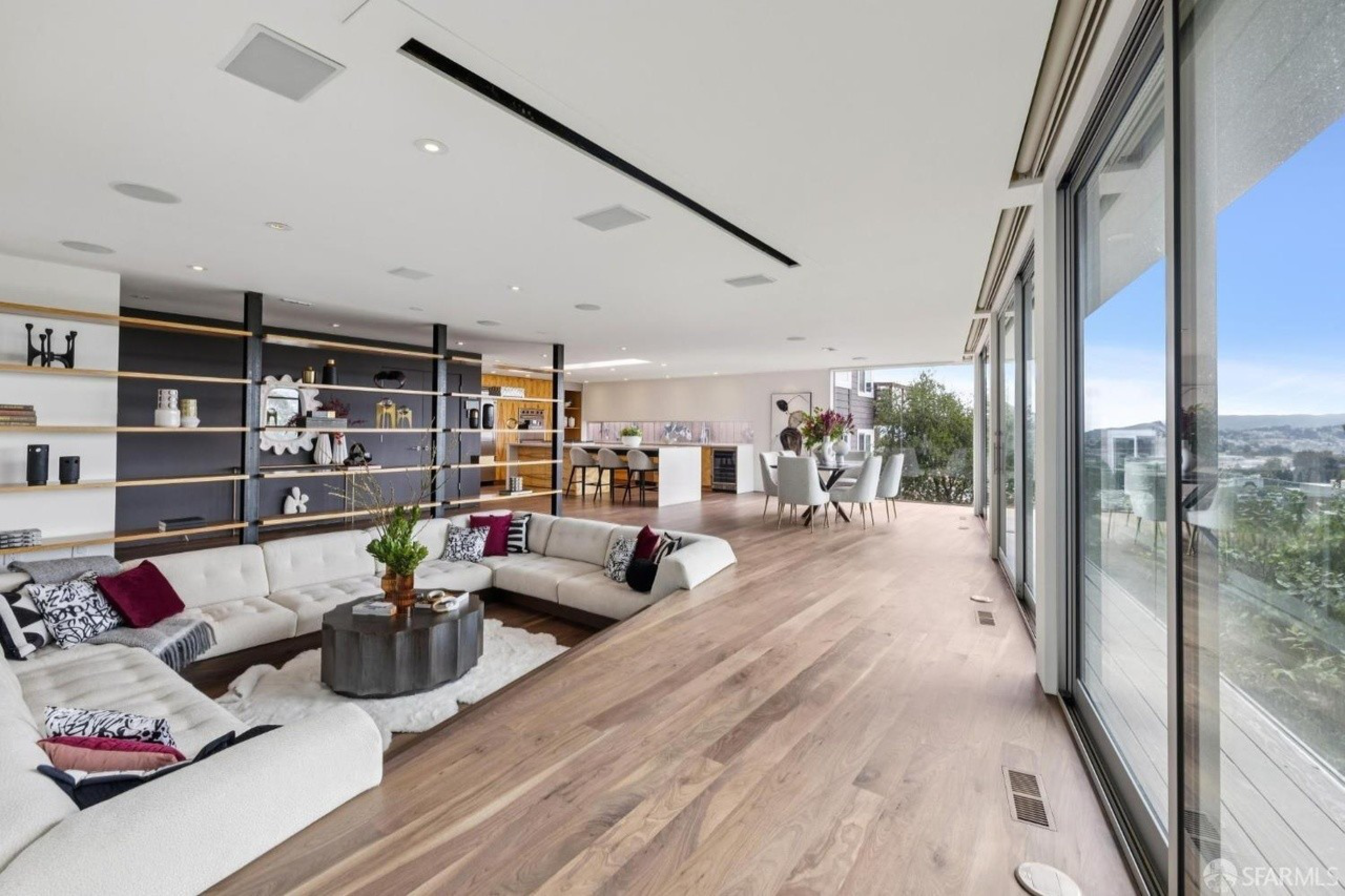 A spacious living area features a large white sectional sofa, round coffee table, open shelves, dining table with chairs, and floor-to-ceiling windows overlooking a cityscape.