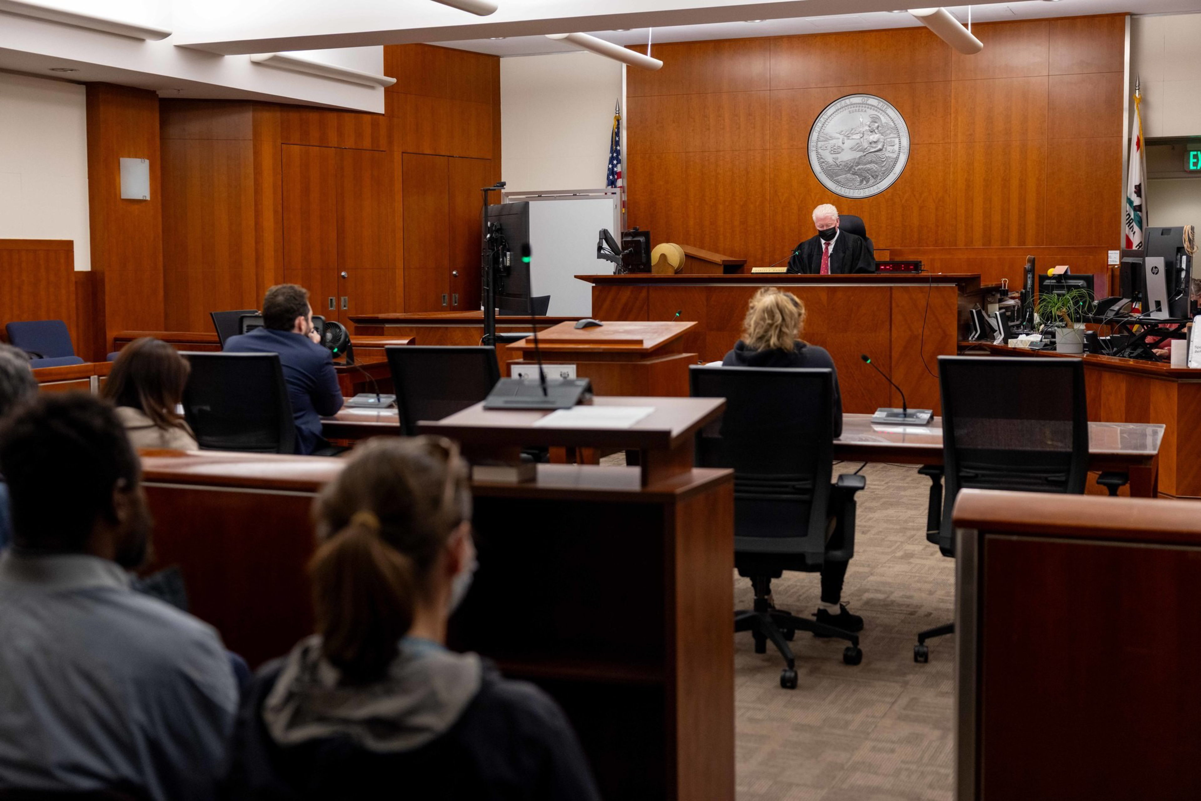 A judge wearing a mask sits at a bench in a wood-paneled courtroom while several people sit in chairs facing toward him.