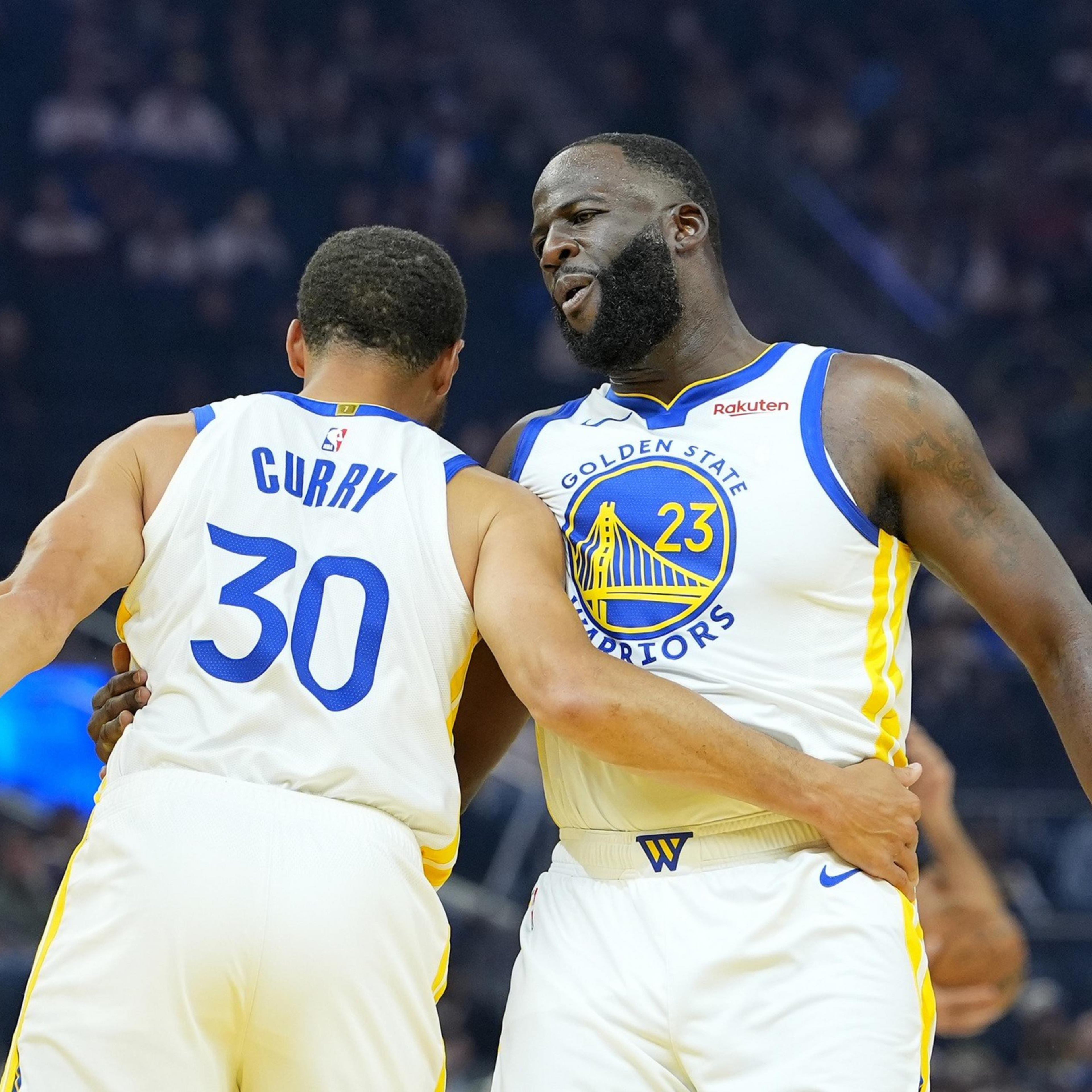 Two Golden State Warriors players, wearing white jerseys with blue and yellow details, embrace during a basketball game.