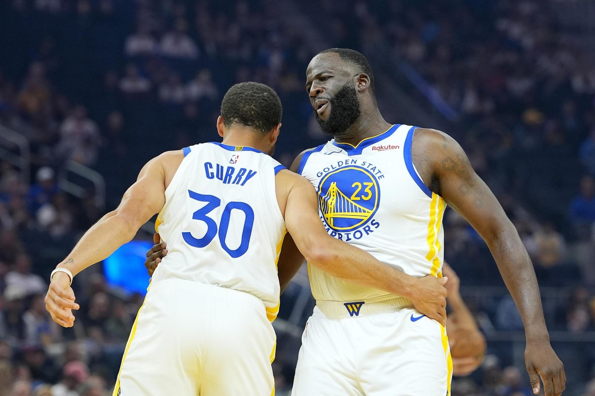 Two Golden State Warriors players, wearing white jerseys with blue and yellow details, embrace during a basketball game.