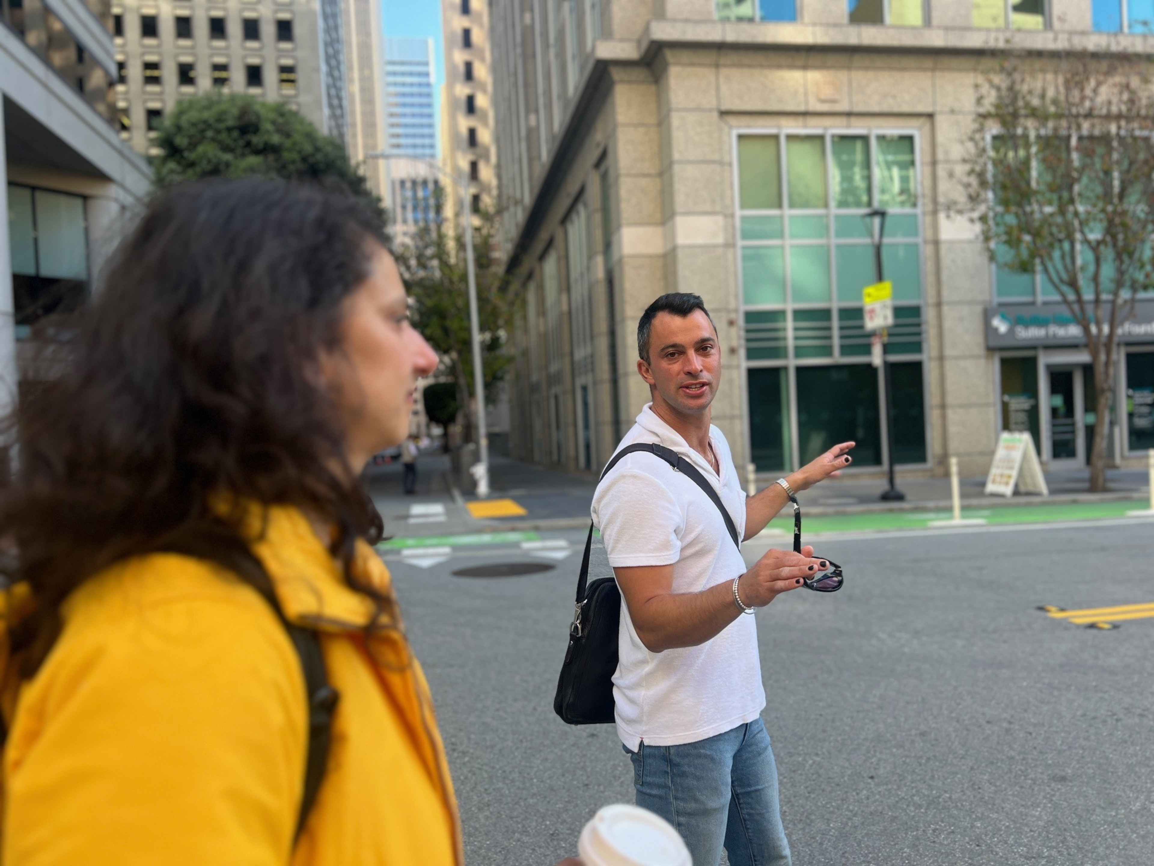 A man holding sunglasses stands in a city street, with a blurred woman in the foreground.