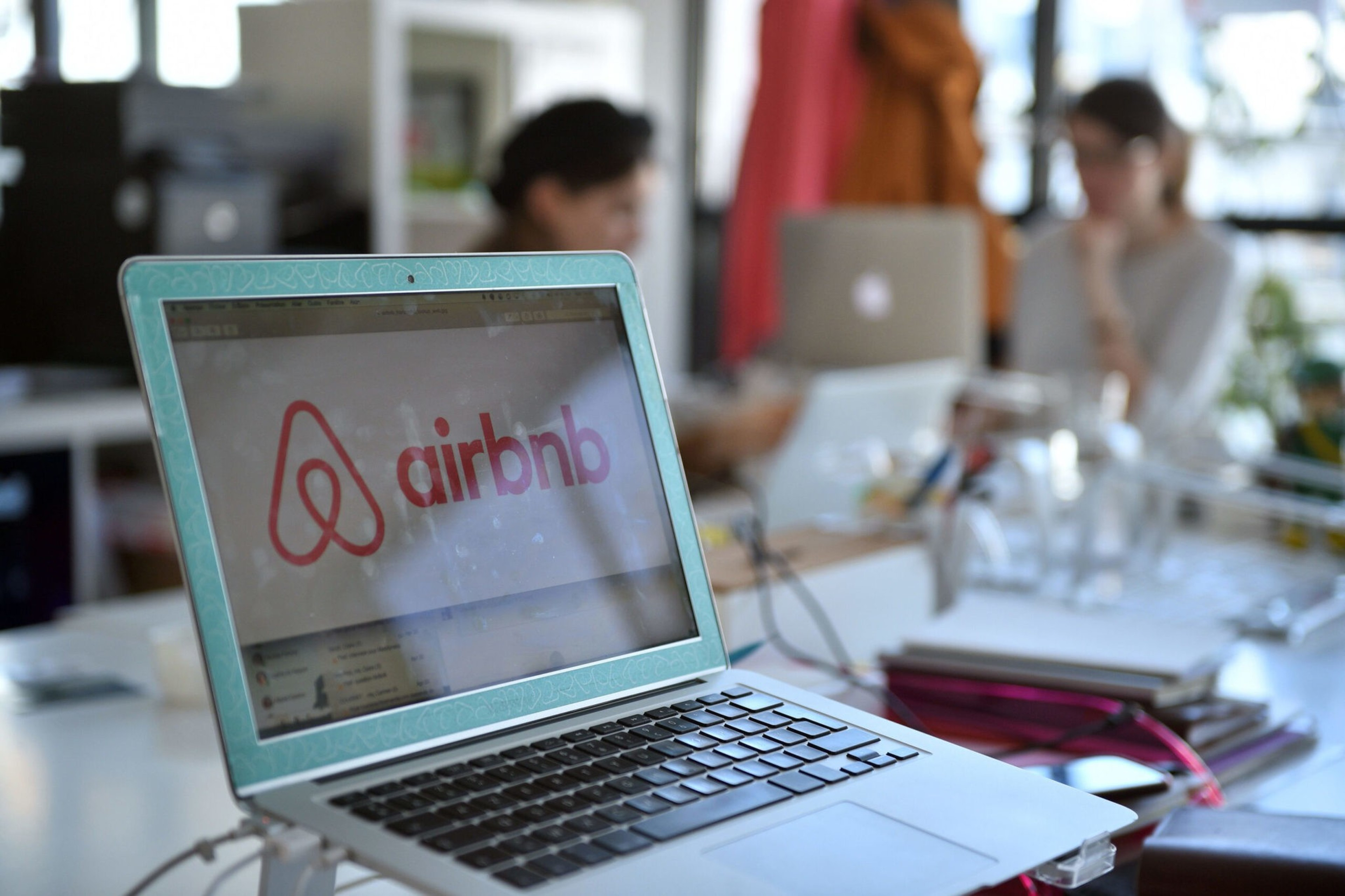 A laptop displays the Airbnb logo on its screen, with two blurred people working in the background at a cluttered desk.