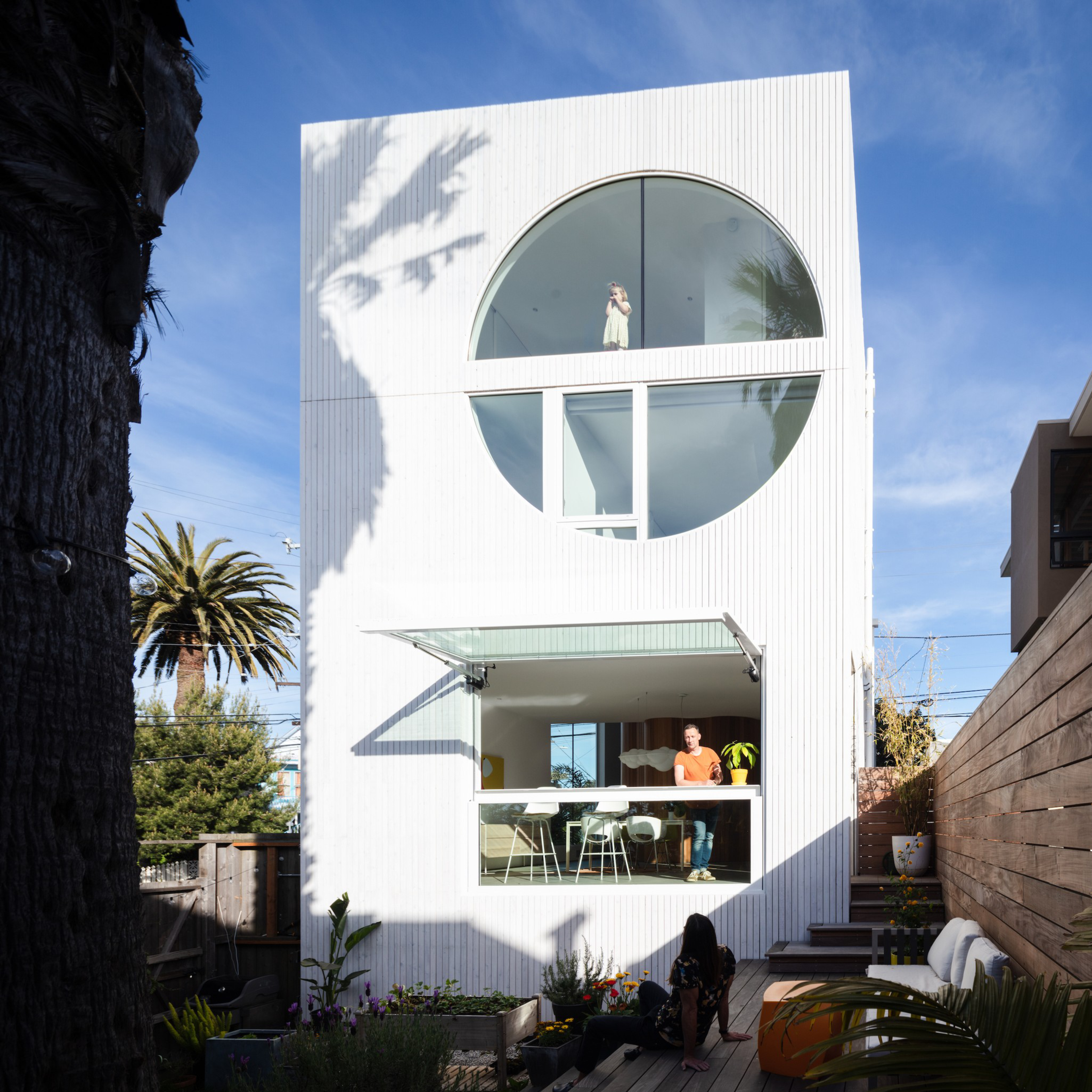 A modern white building with a large circular and a rectangular window. A man stands in the latter; a woman in a dress is visible in the circle. Trees surround the area.