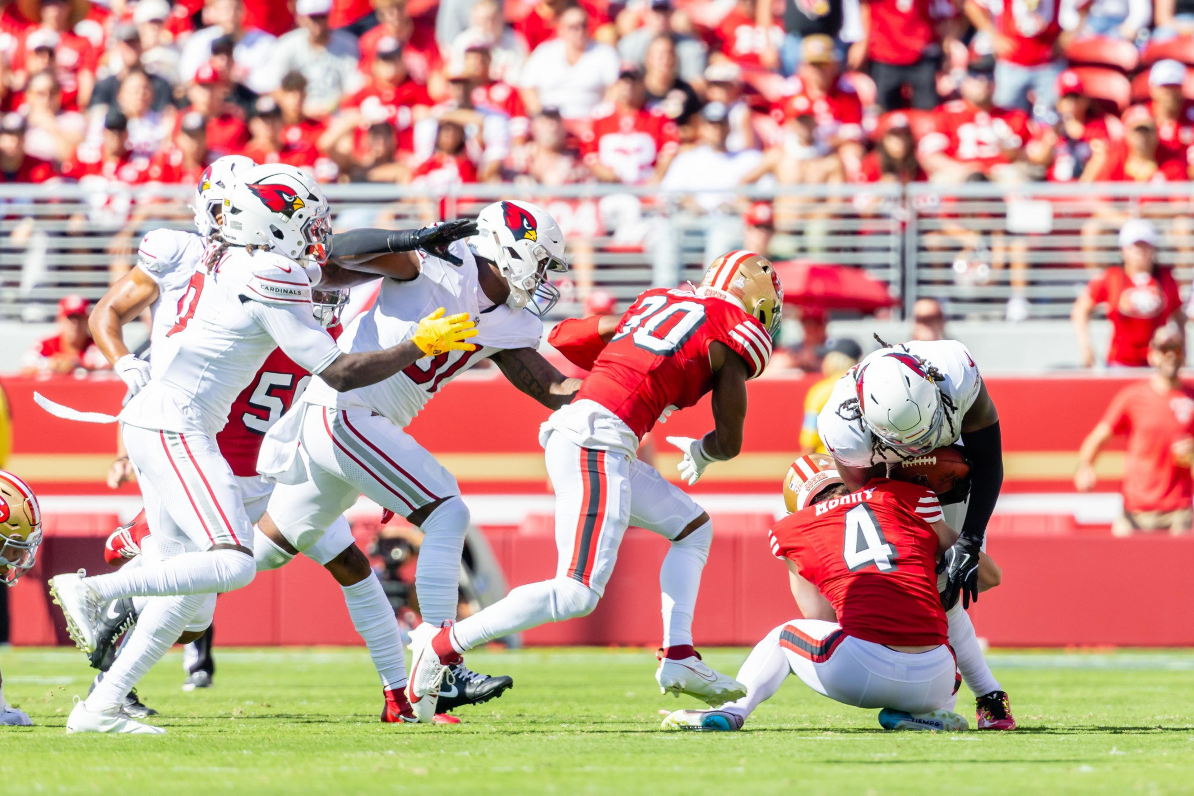 A group of football players in red and white uniforms are actively competing on the field. One player is tackling another who is holding the ball.