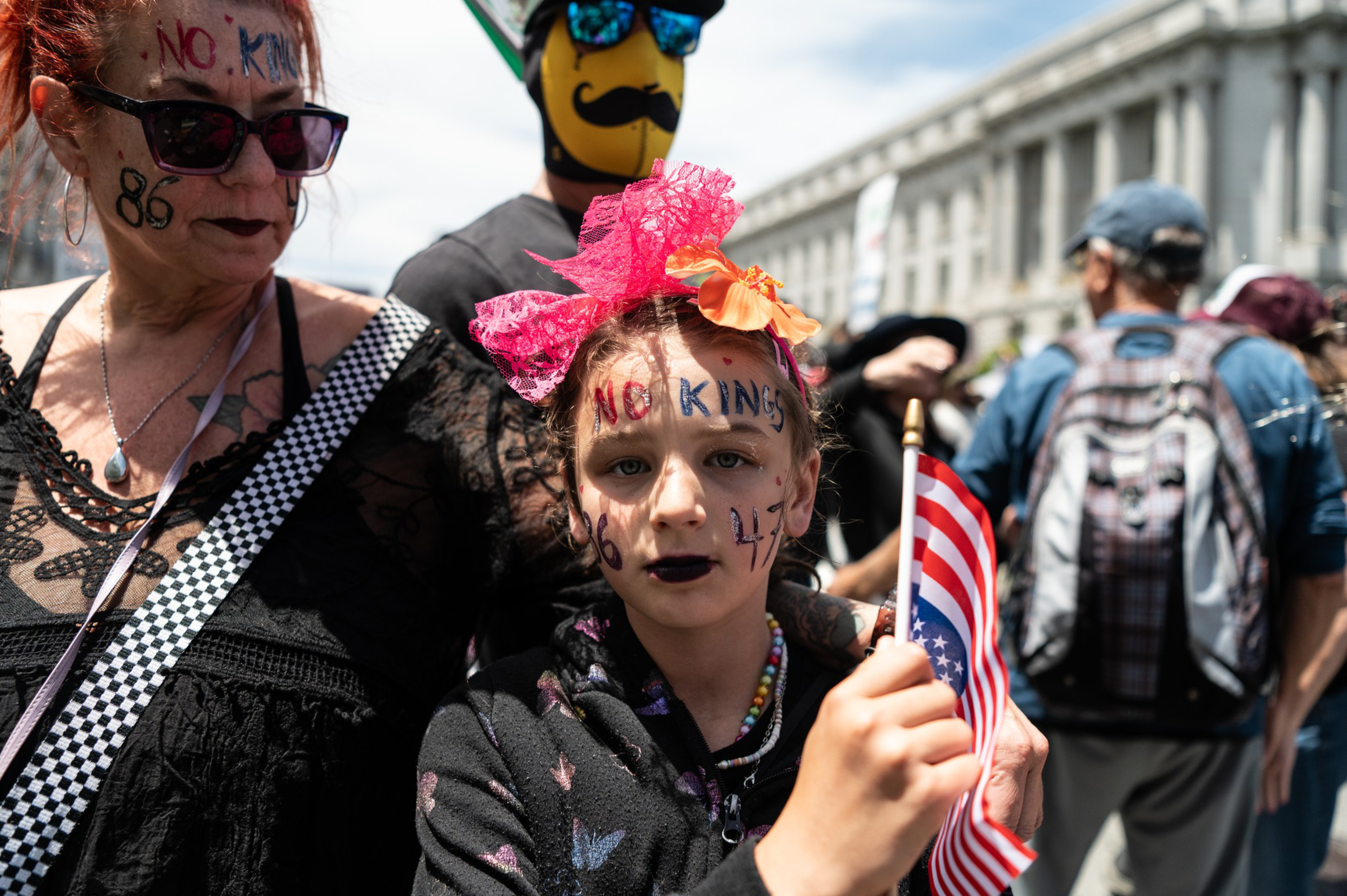 A child holds a flag, wearing face paint that says &quot;No King,&quot; with bright decorations in their hair. They stand among a crowd, including a person in sunglasses.
