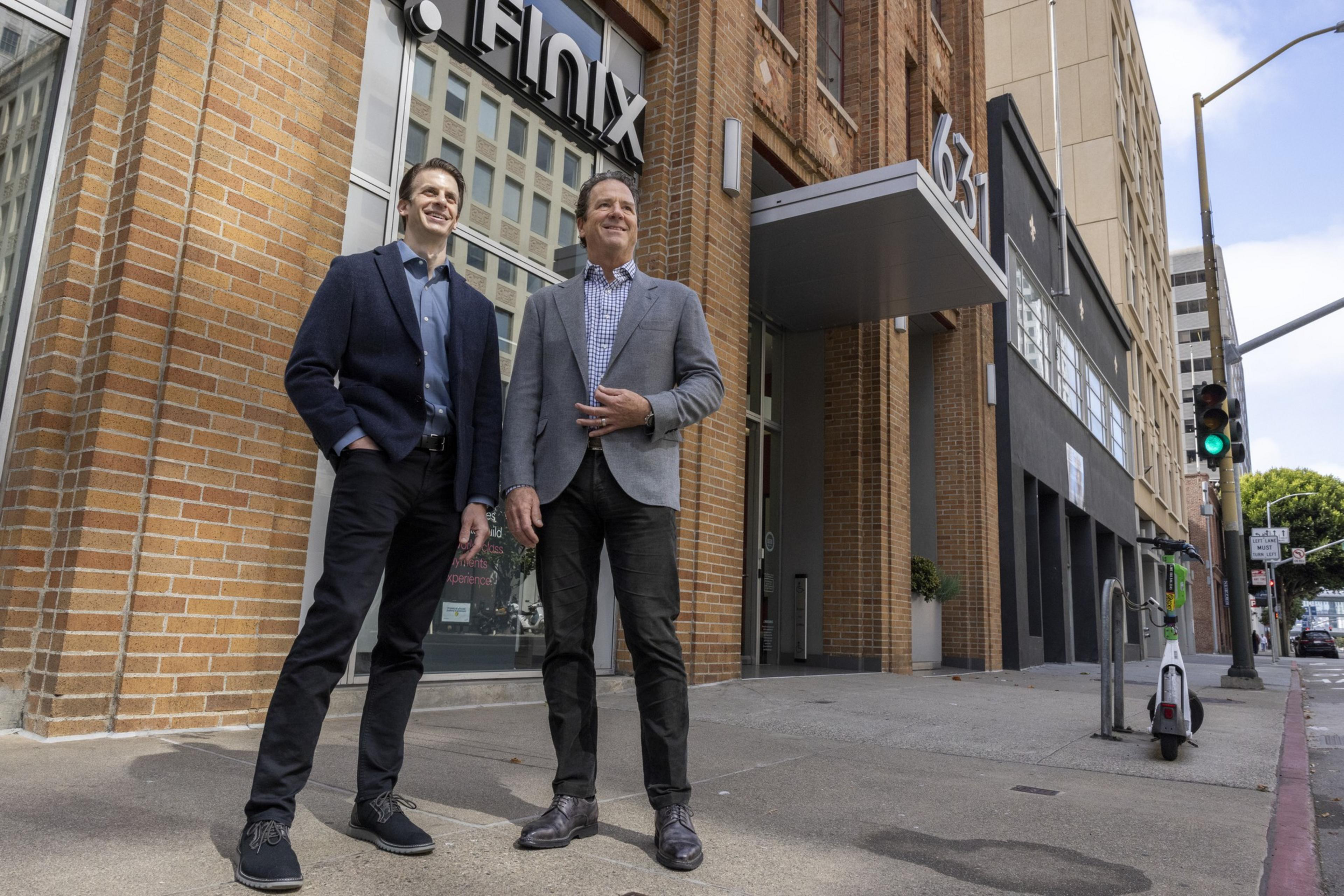 Two men in business attire stand outside a brick building labeled "Finix" with a street address of 631. The street beside them has a green traffic light and an electric scooter.