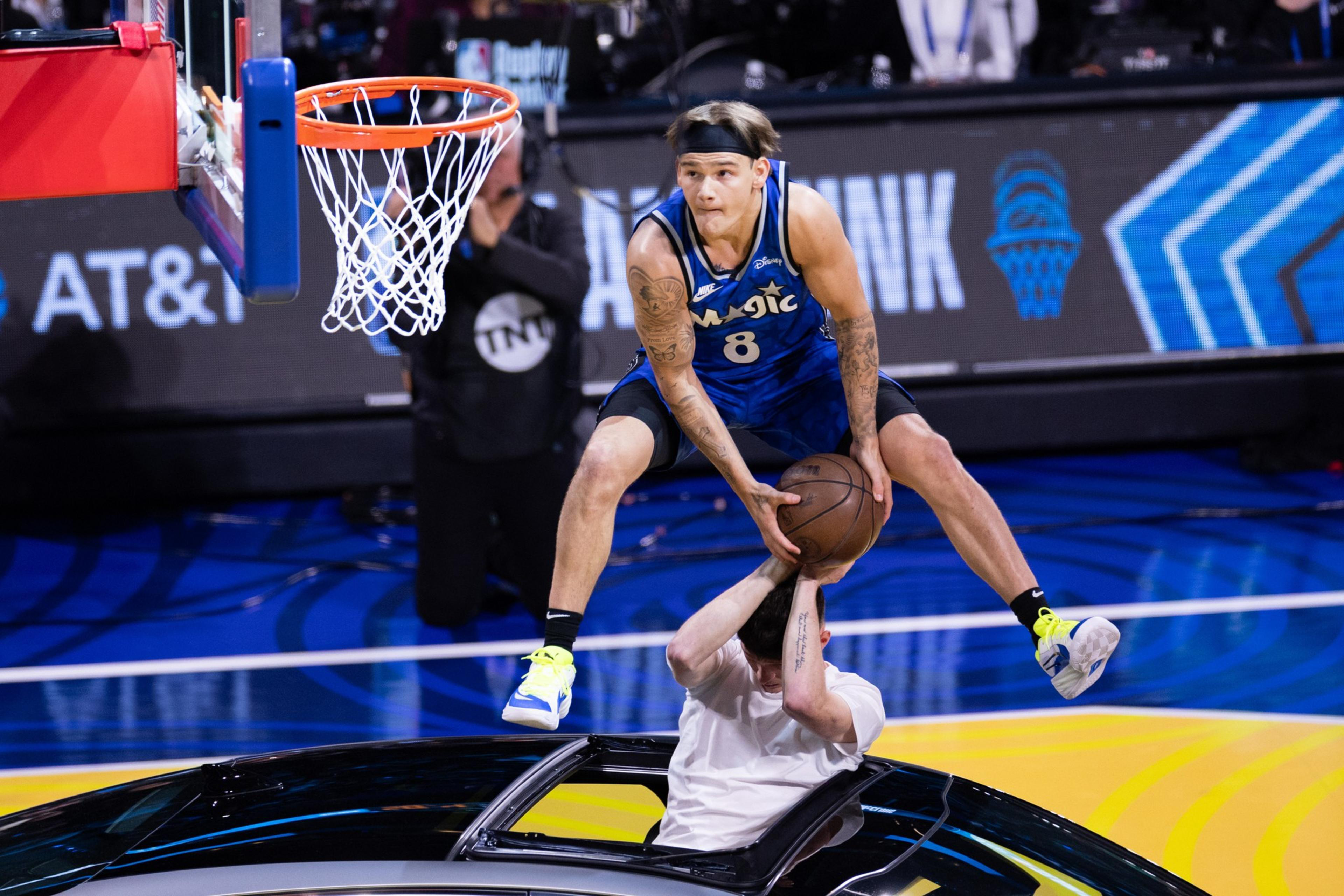 A basketball player in a blue jersey jumps over a person emerging from a car sunroof, holding a basketball, near a hoop.