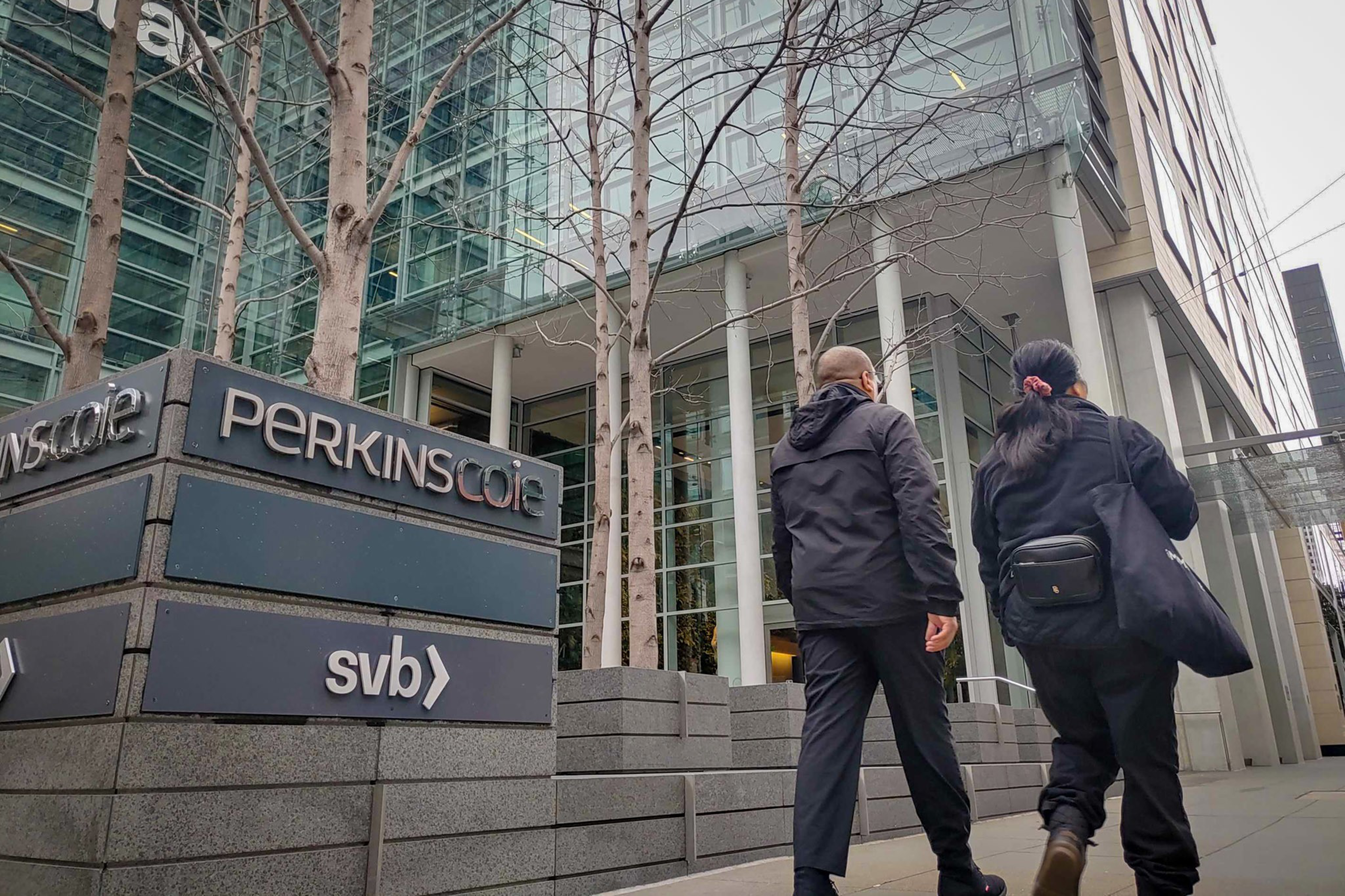 Two people walk past signs for "Perkins Coie" and "svb" outside a modern glass building with leafless trees lining the sidewalk.