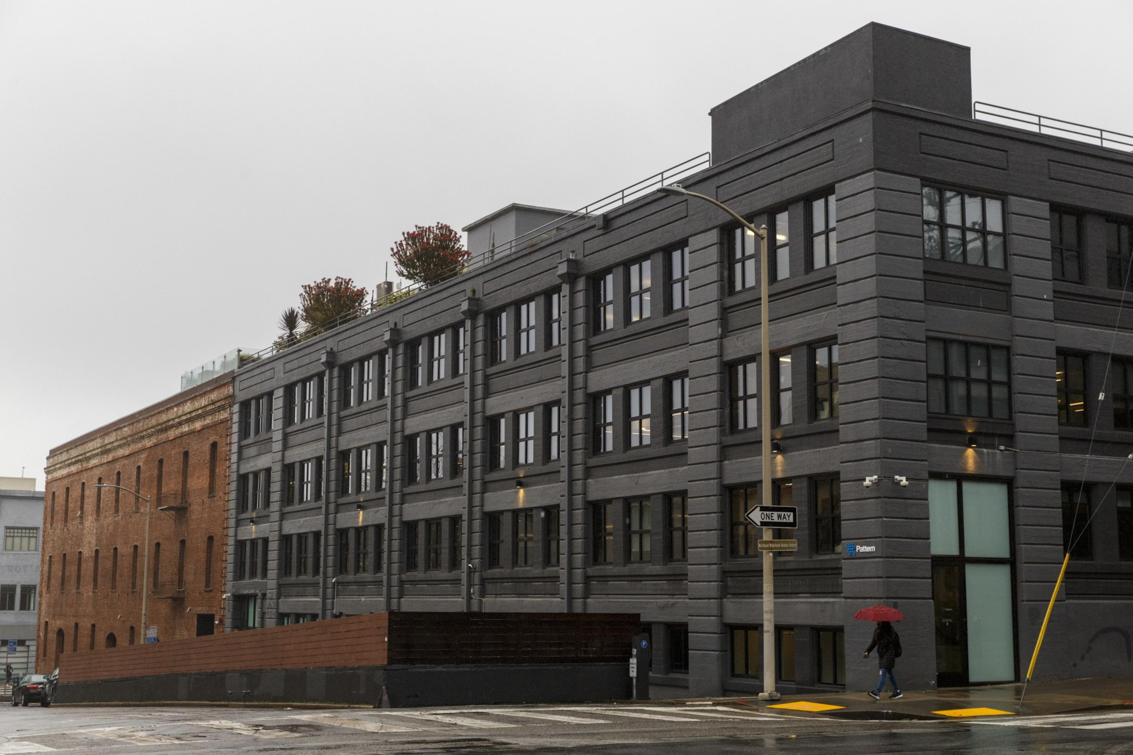 A modern gray building next to an older brick one on a wet street, with a person holding a red umbrella walking by.