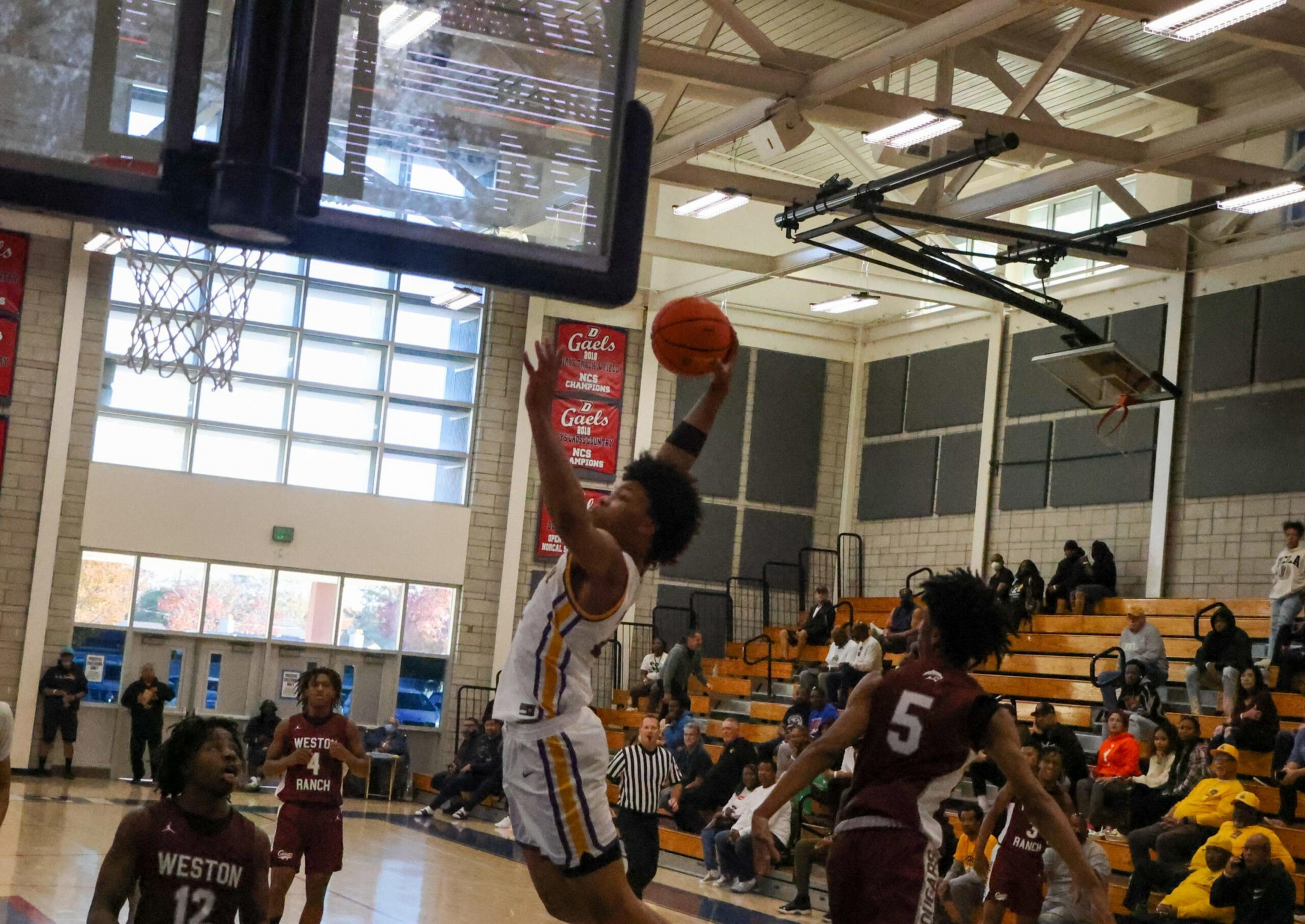 A basketball player in white jumps to score while two players in maroon jerseys defend under an indoor gym with spectators seated in bleachers.
