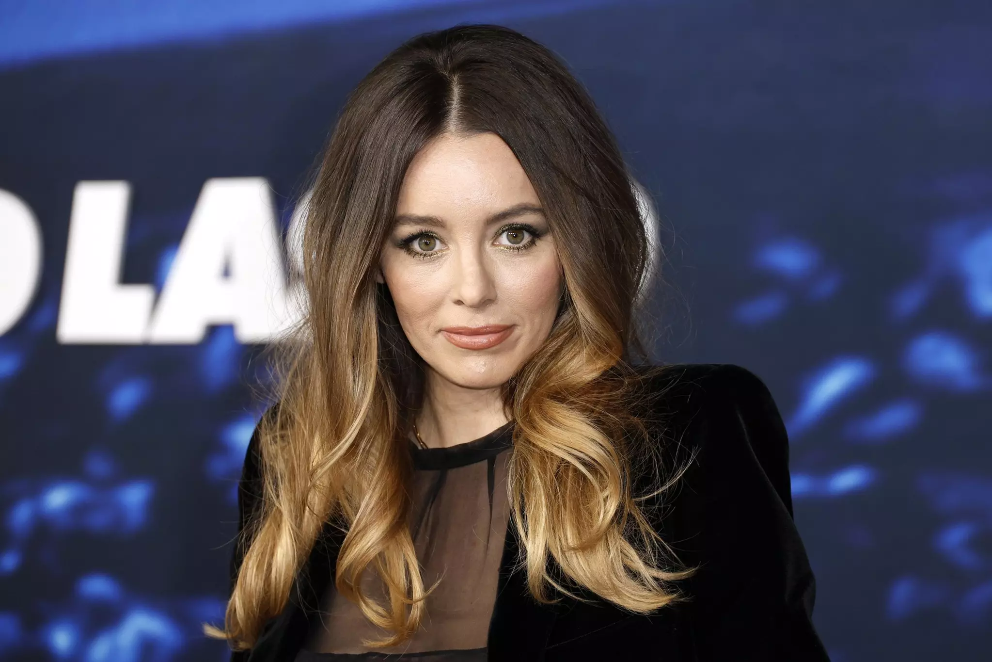 A woman with long, wavy brown hair with blonde ends wears a black outfit with a sheer top, looking directly at the camera against a dark blue background.