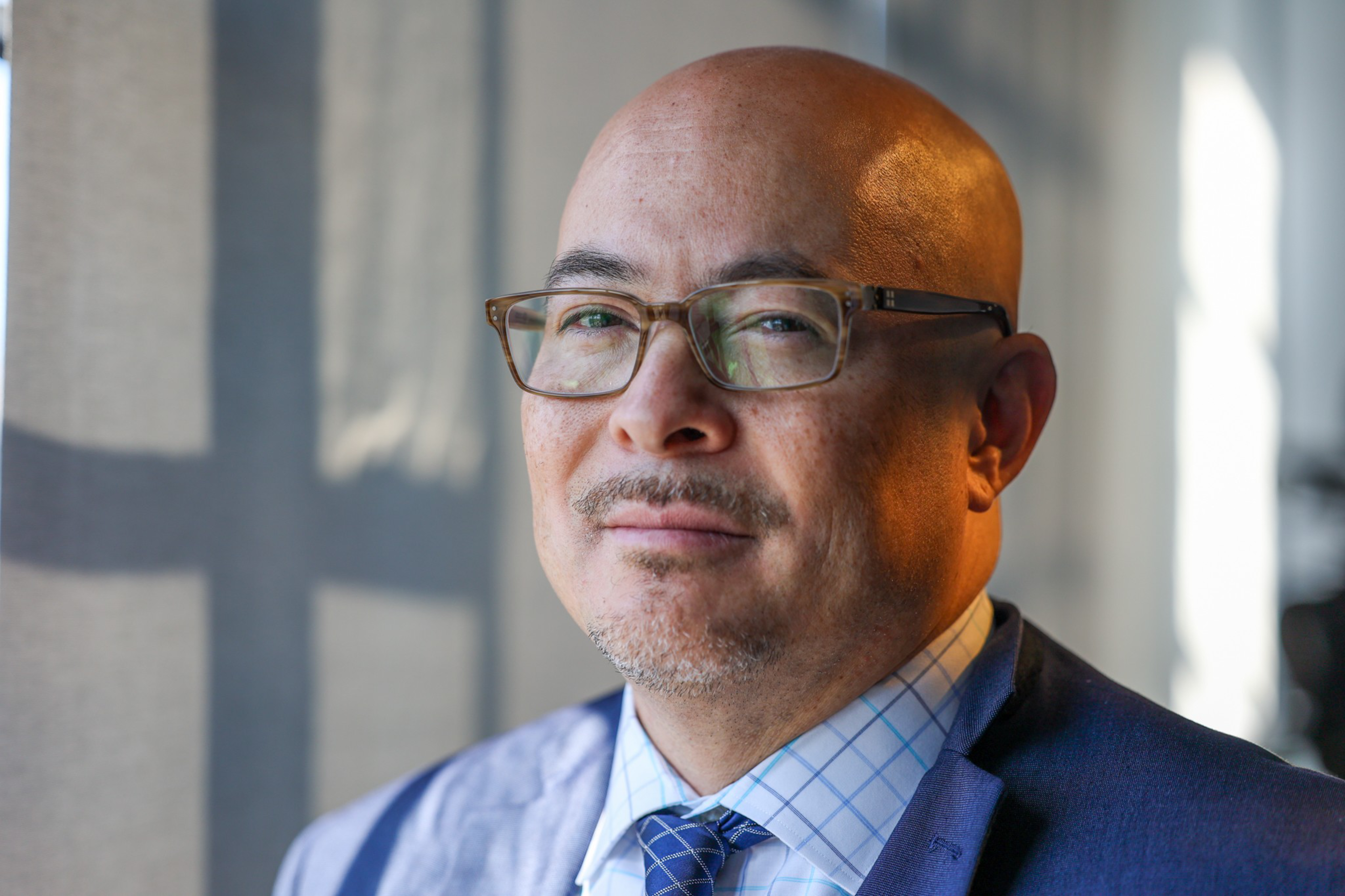 A middle-aged bald man with glasses and a slight mustache wears a blue suit, checkered shirt, and blue tie, looking calmly at the camera in soft natural light.