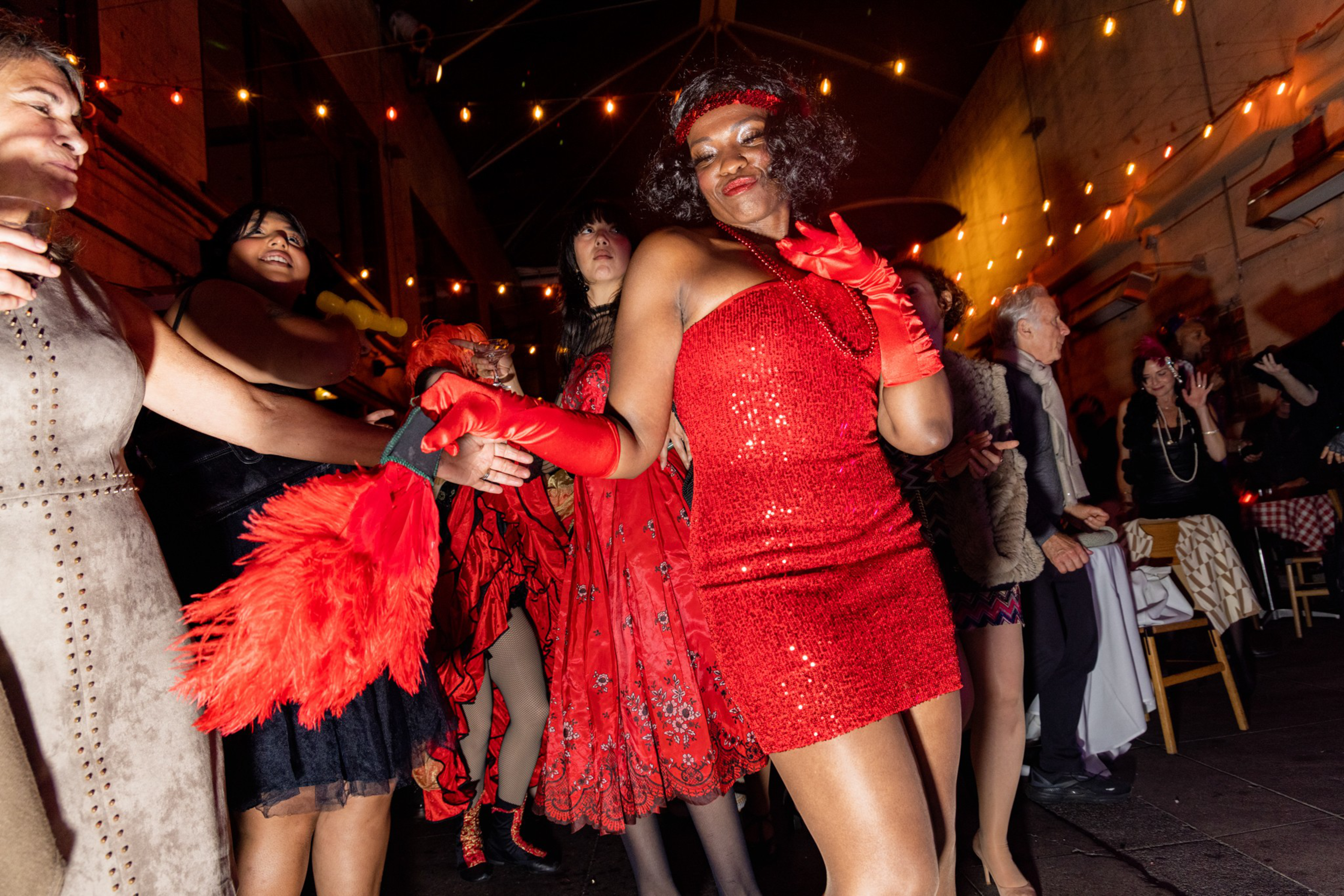 A lively group of people dances under string lights, with one woman in a sparkly red dress and matching gloves standing out in the center.