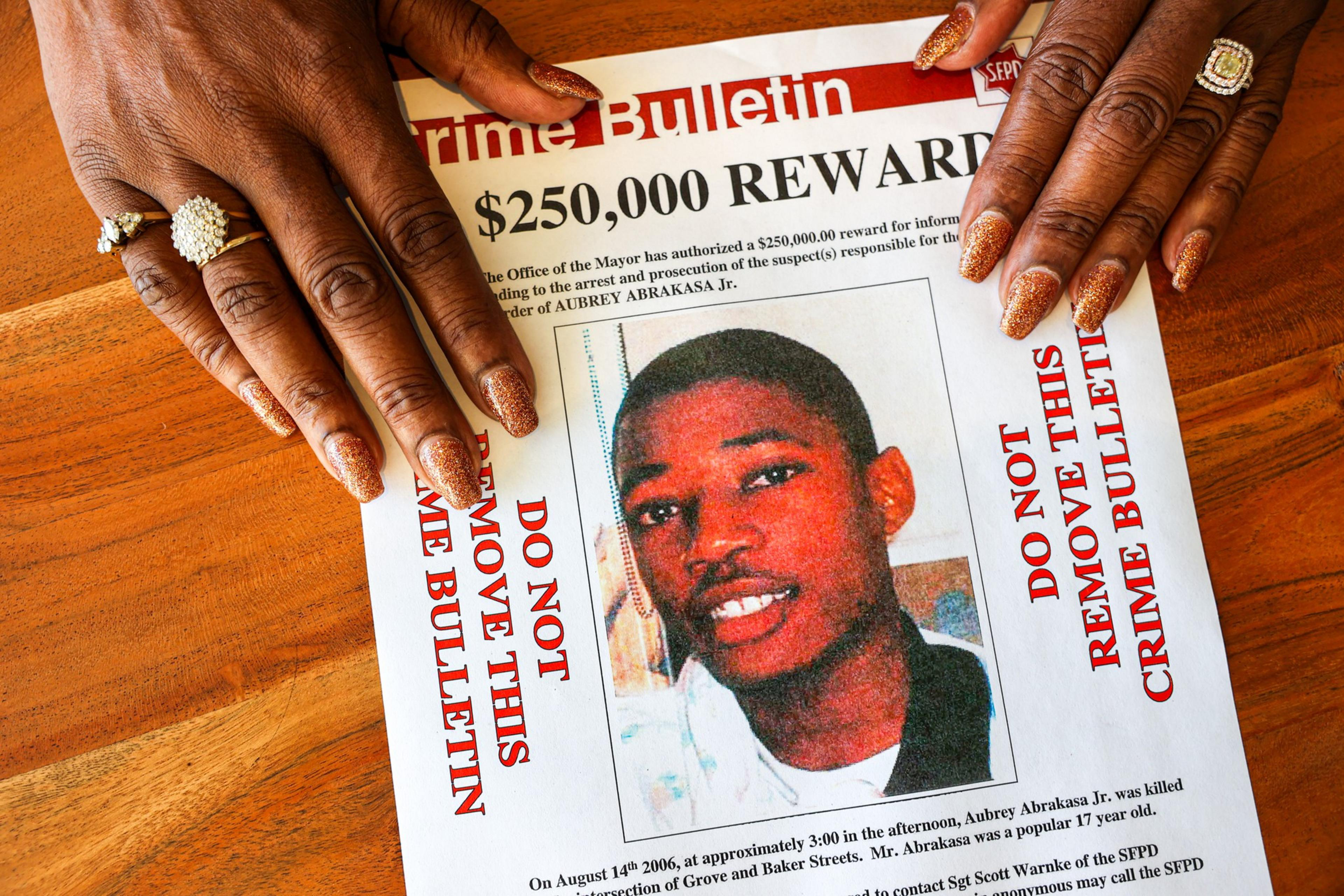 A close-up of a crime bulletin flyer on a wooden table, featuring a $250,000 reward for information. Two hands with decorated nails frame the flyer.