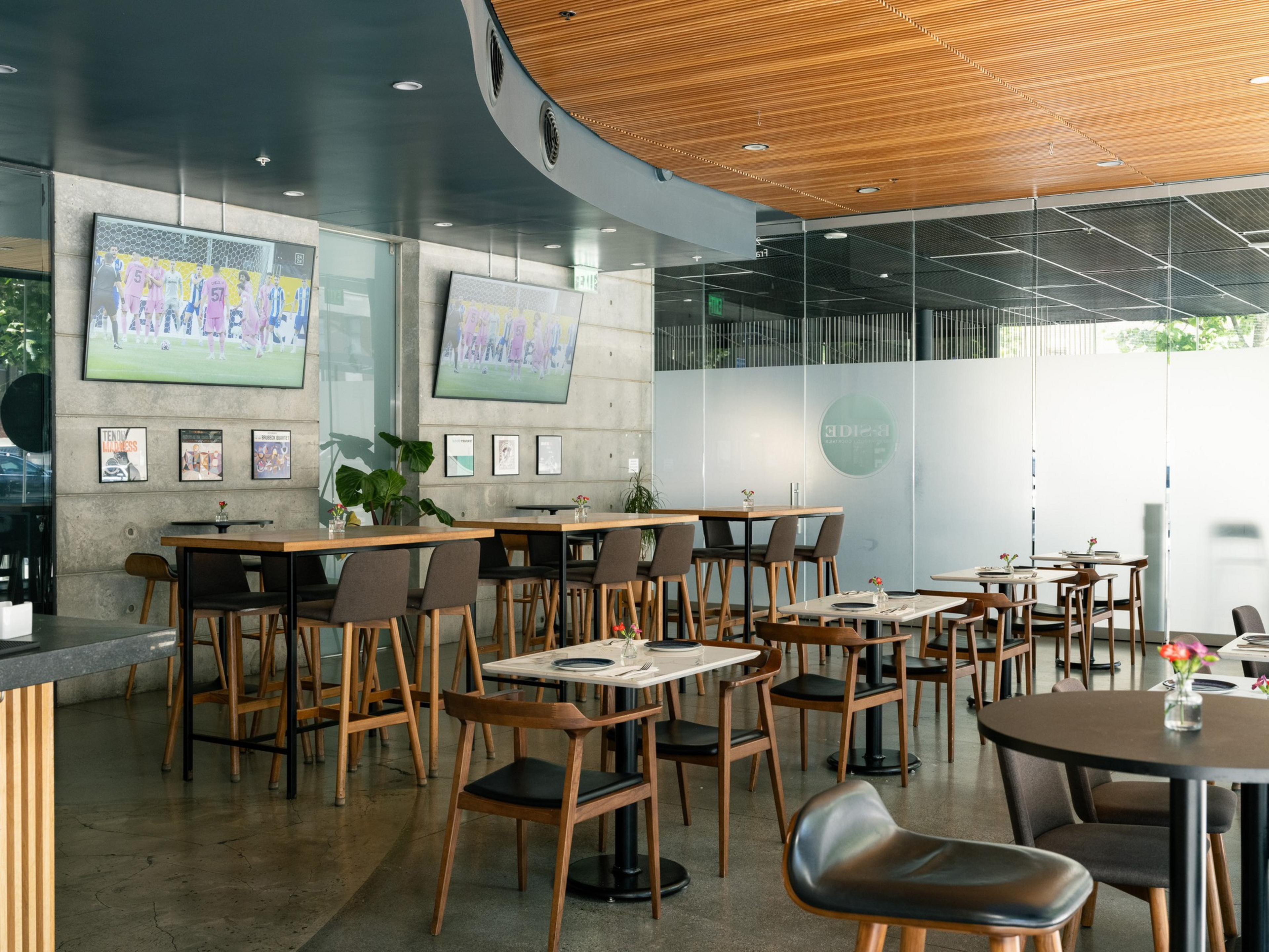 The image shows a modern cafe with wooden tables and chairs, two TVs on the wall displaying a soccer game, framed pictures, and a glass partition.