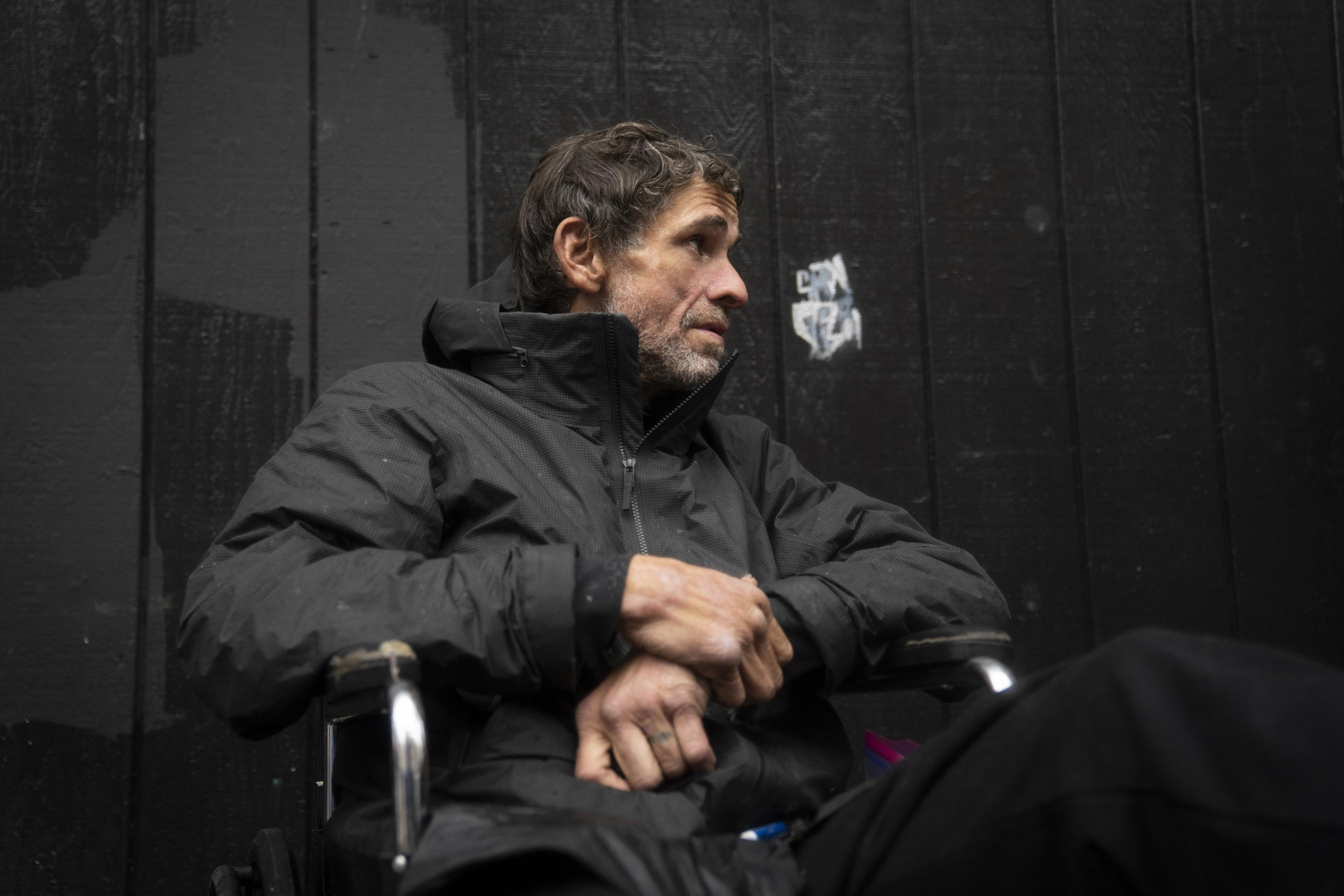 A man in a wheelchair, wearing a black jacket, sits pensively against a dark wooden wall. His hands are clasped together, and he gazes thoughtfully to the side.