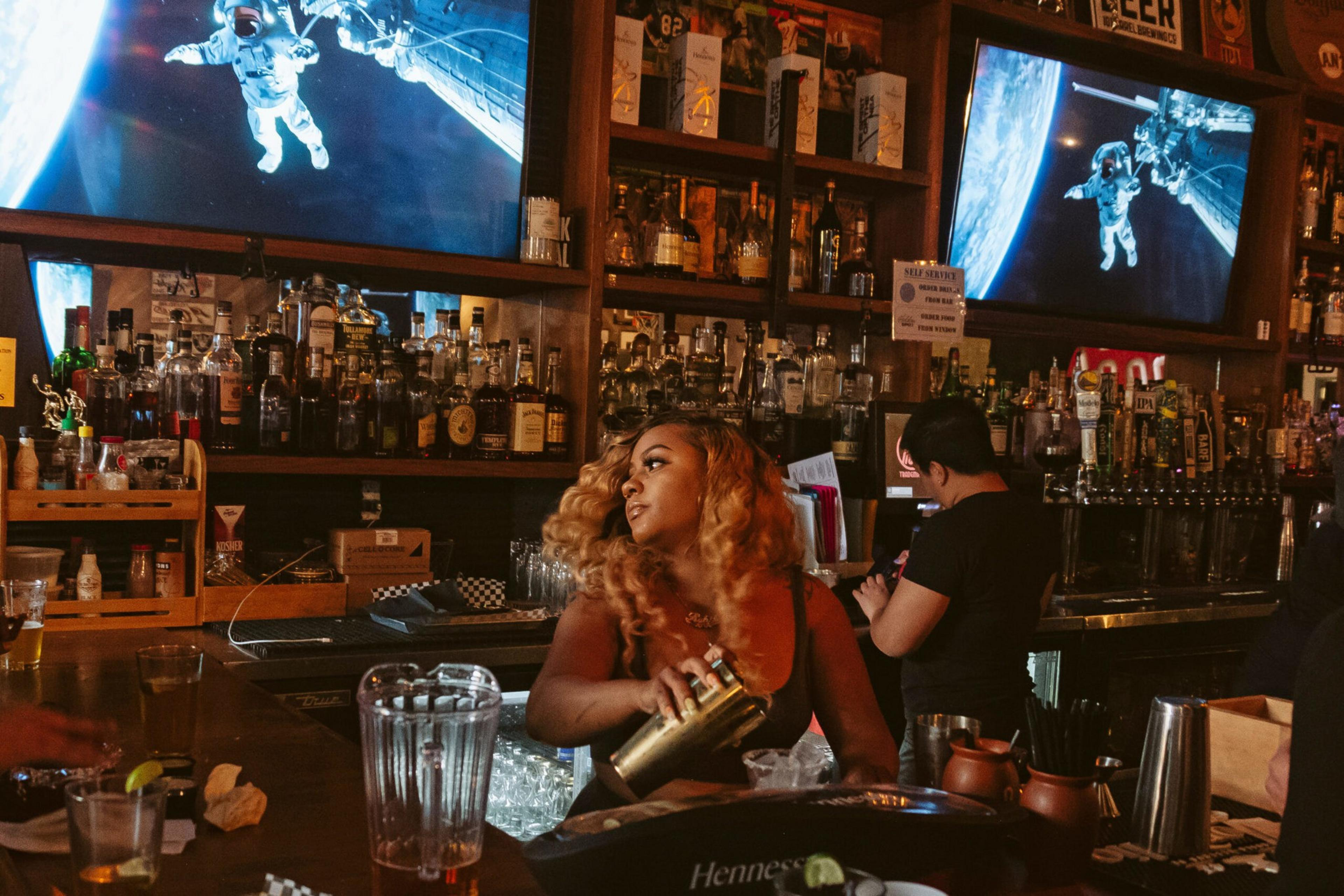 A bartender with curly hair shakes a cocktail shaker behind a busy bar, with an astronaut scene playing on two large screens above.