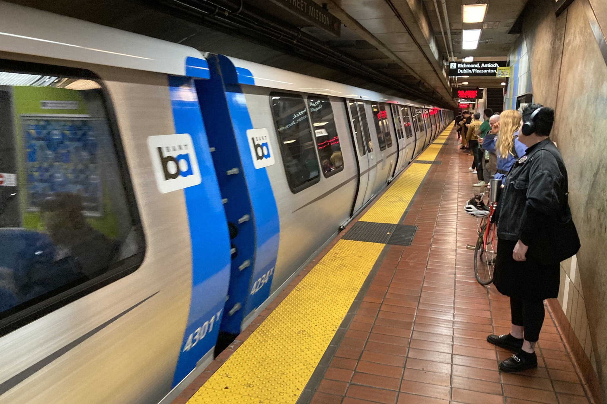 A BART train stands at an underground platform with several passengers waiting, including a person wearing headphones near a bike.