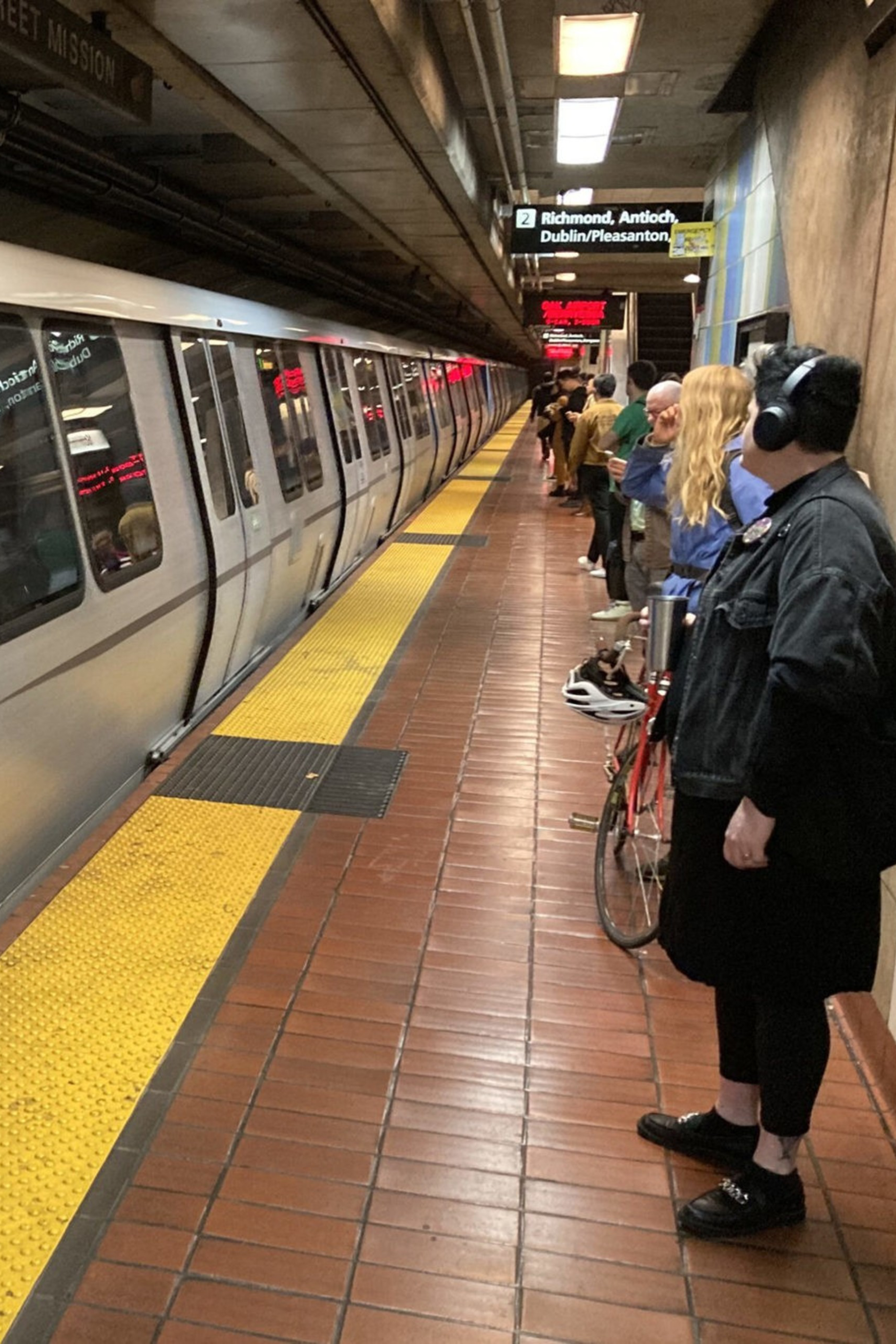 A BART train stands at an underground platform with several passengers waiting, including a person wearing headphones near a bike.
