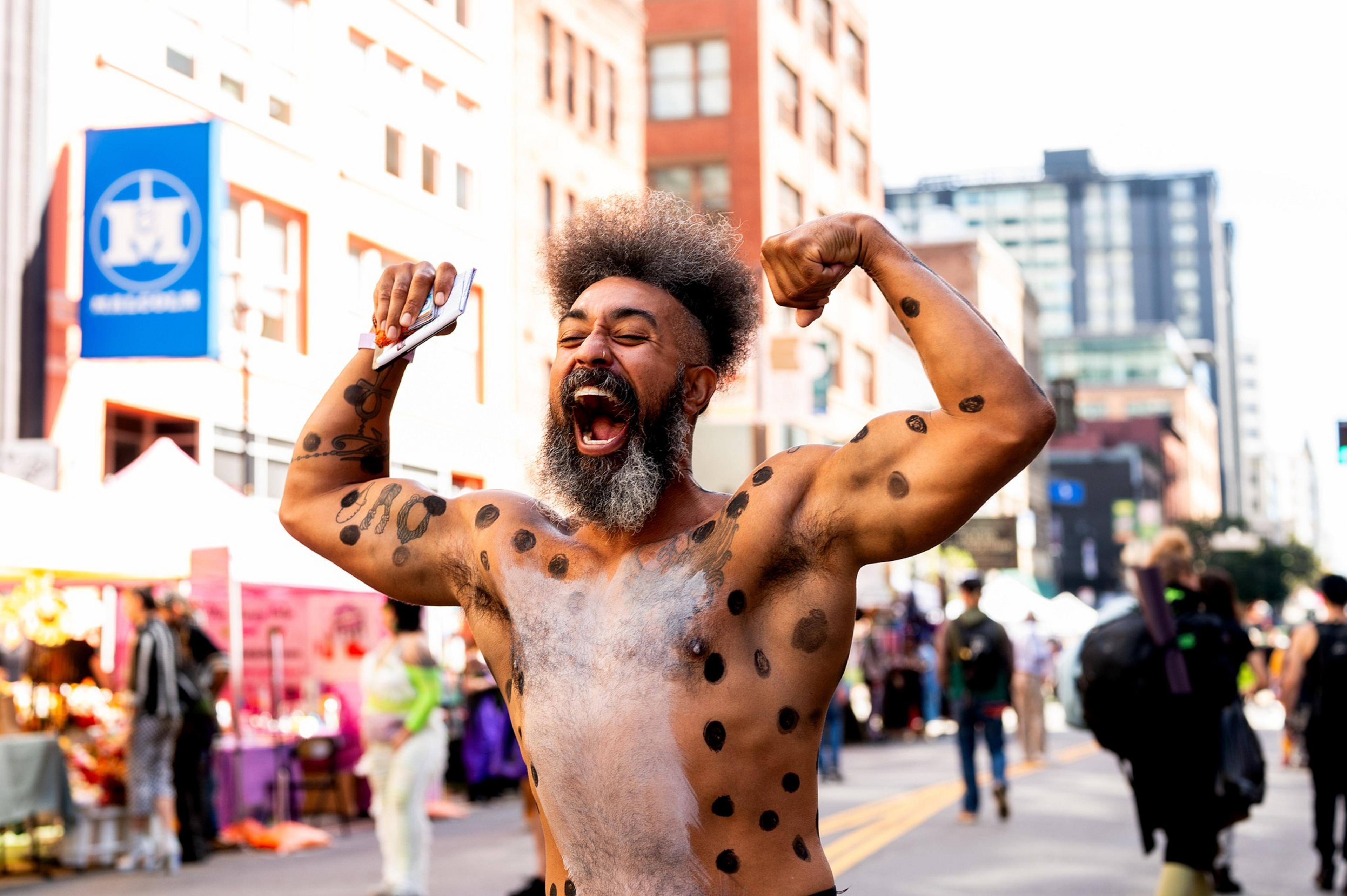 A man with an expressive face flexes his muscles on a busy street, his body painted with black spots. He appears energetic amidst a lively, urban setting.