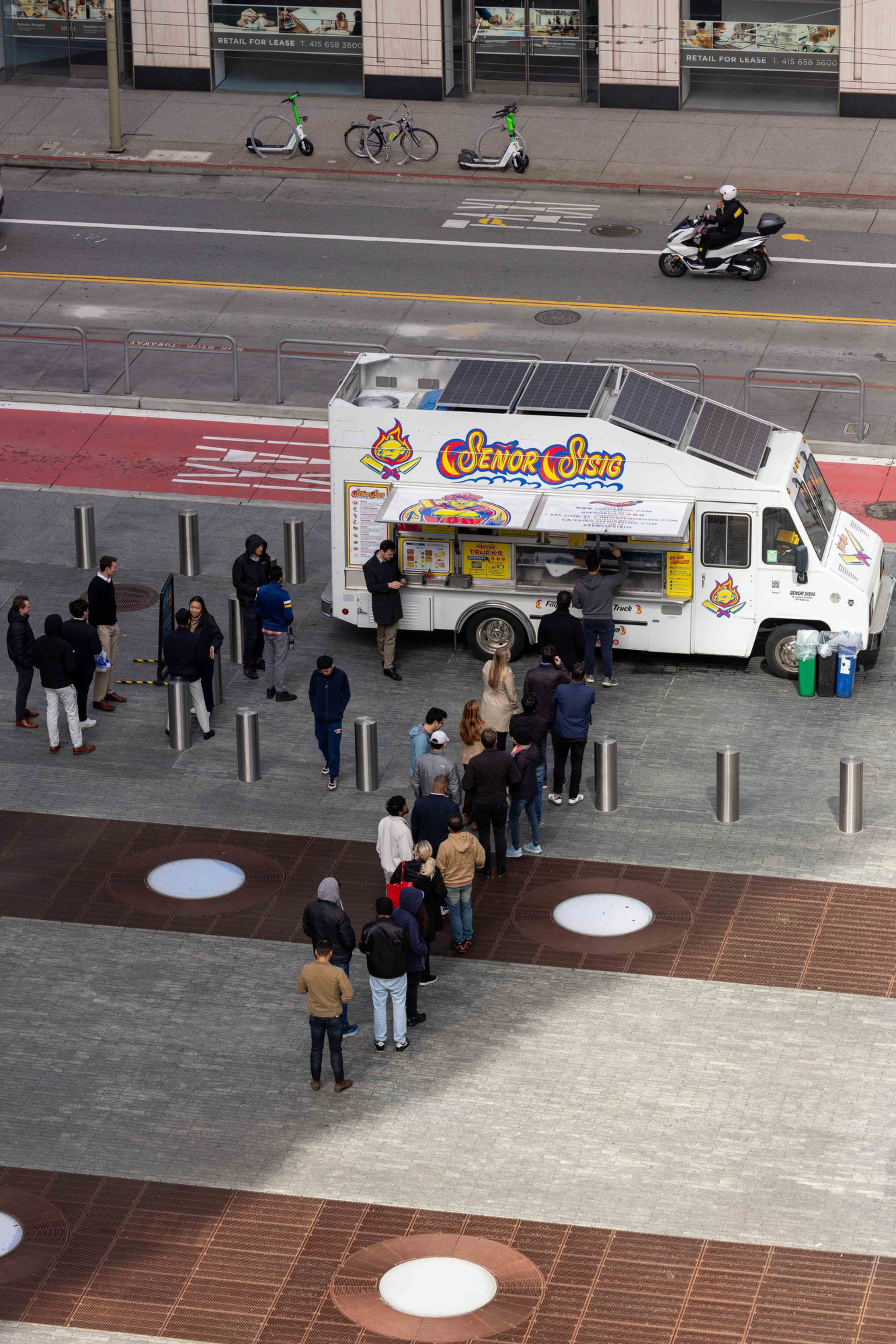 A food truck with "Señor Sisig" branding has a line of people waiting in front. The truck is parked on a paved area by the street, where scooters and a motorcycle are visible.