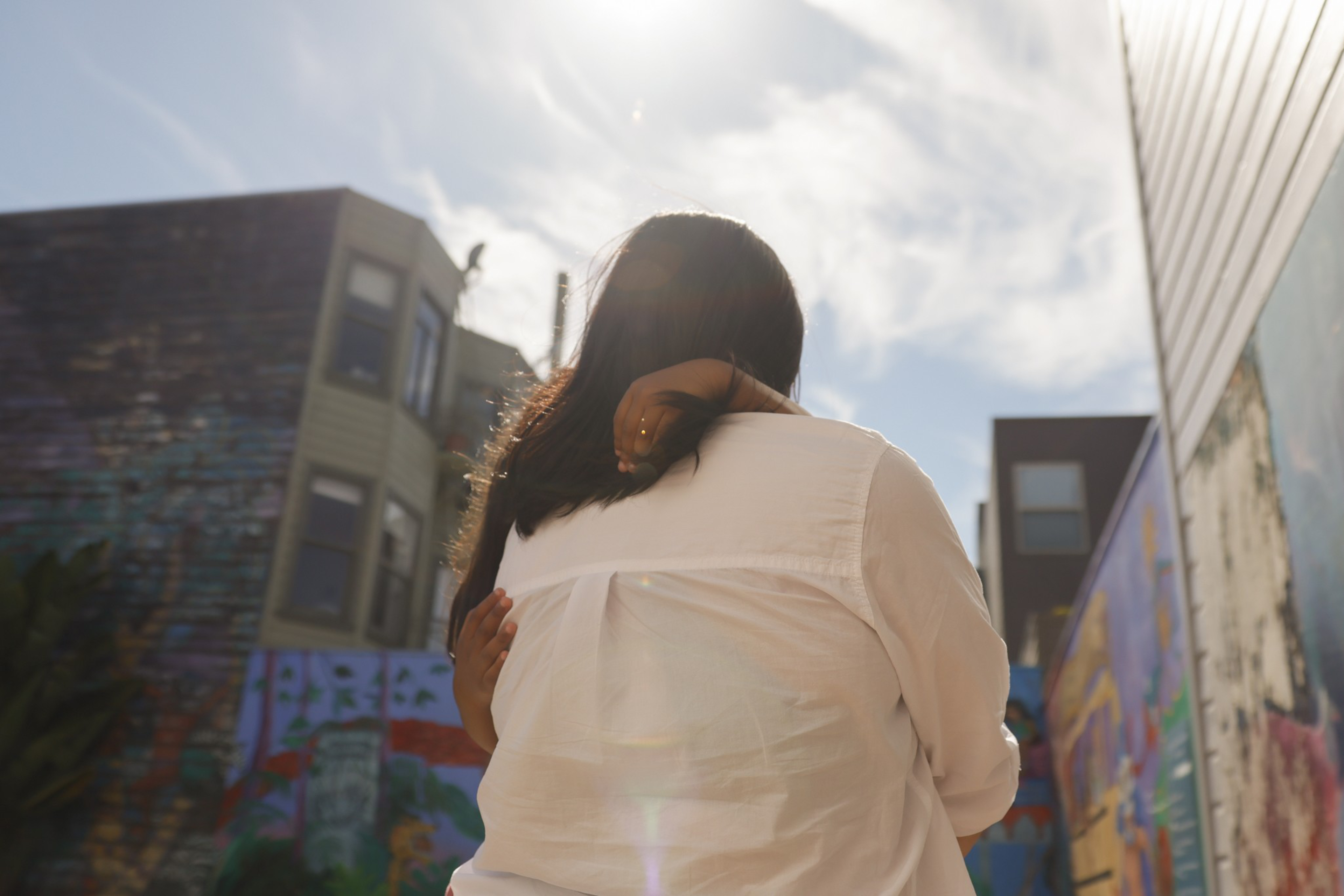 A person wearing a white shirt is hugging a child with long dark hair outdoors, with colorful murals and buildings in the background under a bright sky.