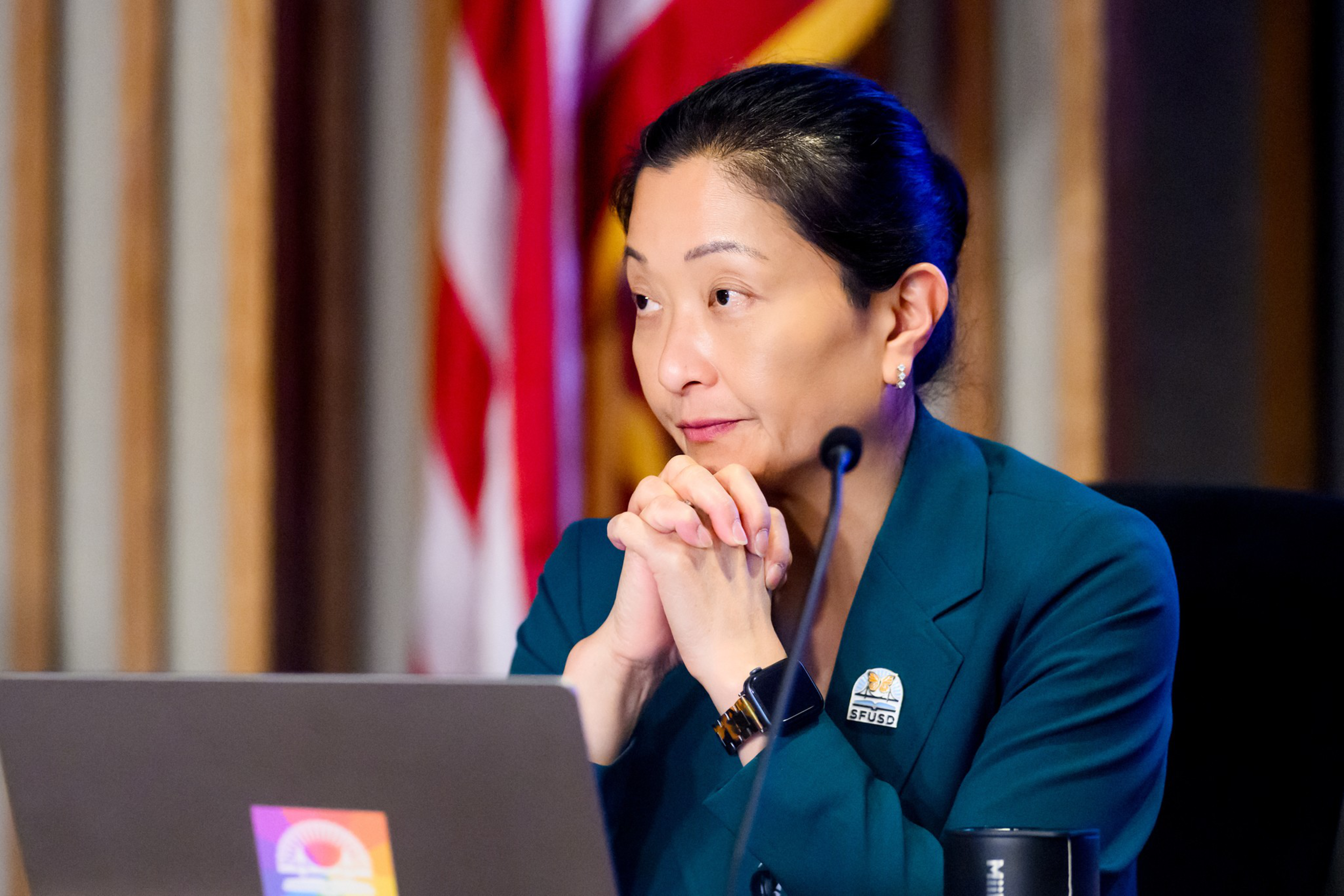 A woman in a teal blazer sits behind a laptop with clasped hands, looking thoughtfully to the side, with a blurred American flag in the background.