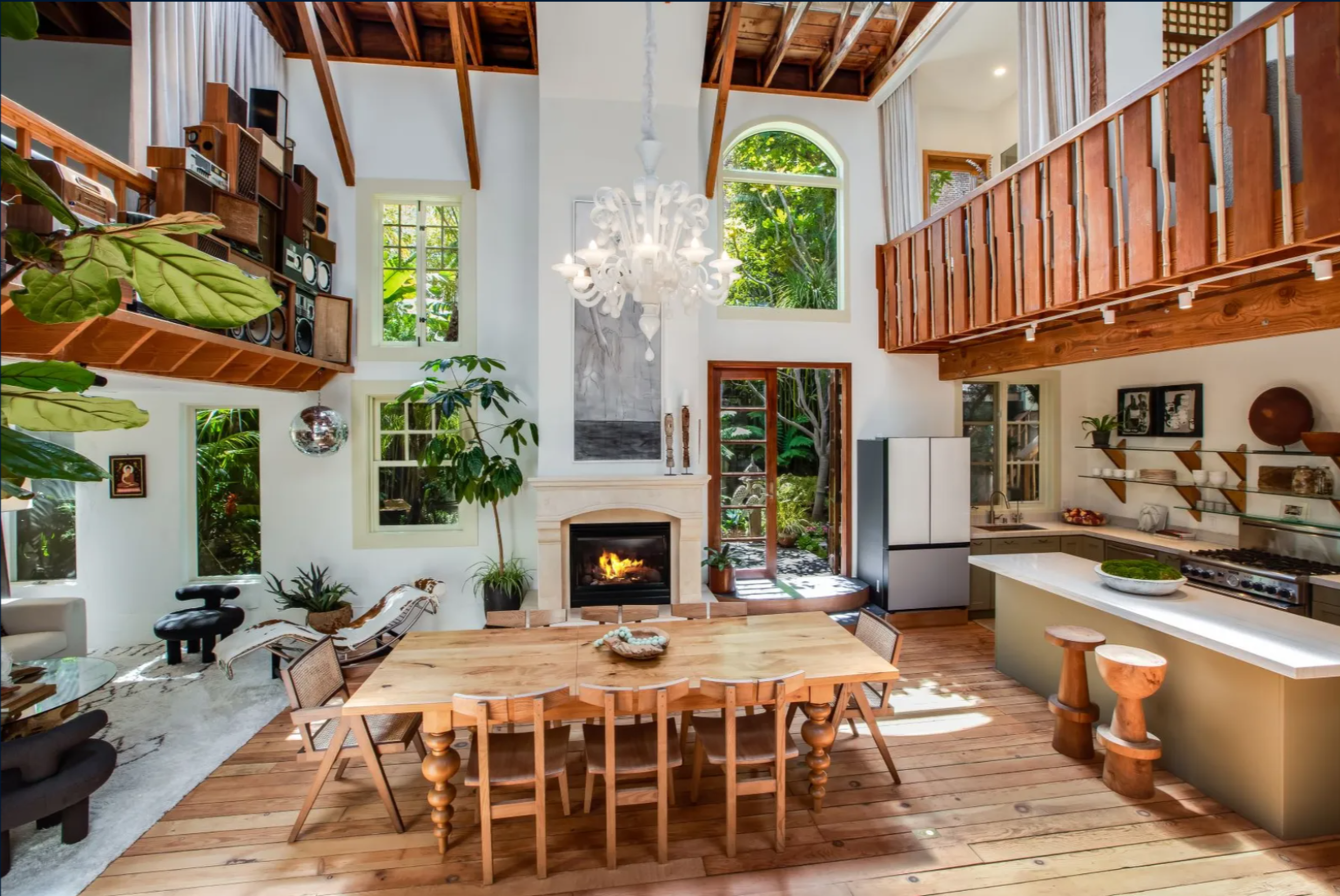 A bright, open living space features wooden floors, a large dining table, a cozy fireplace, indoor plants, a kitchen island with stools, and a mezzanine.