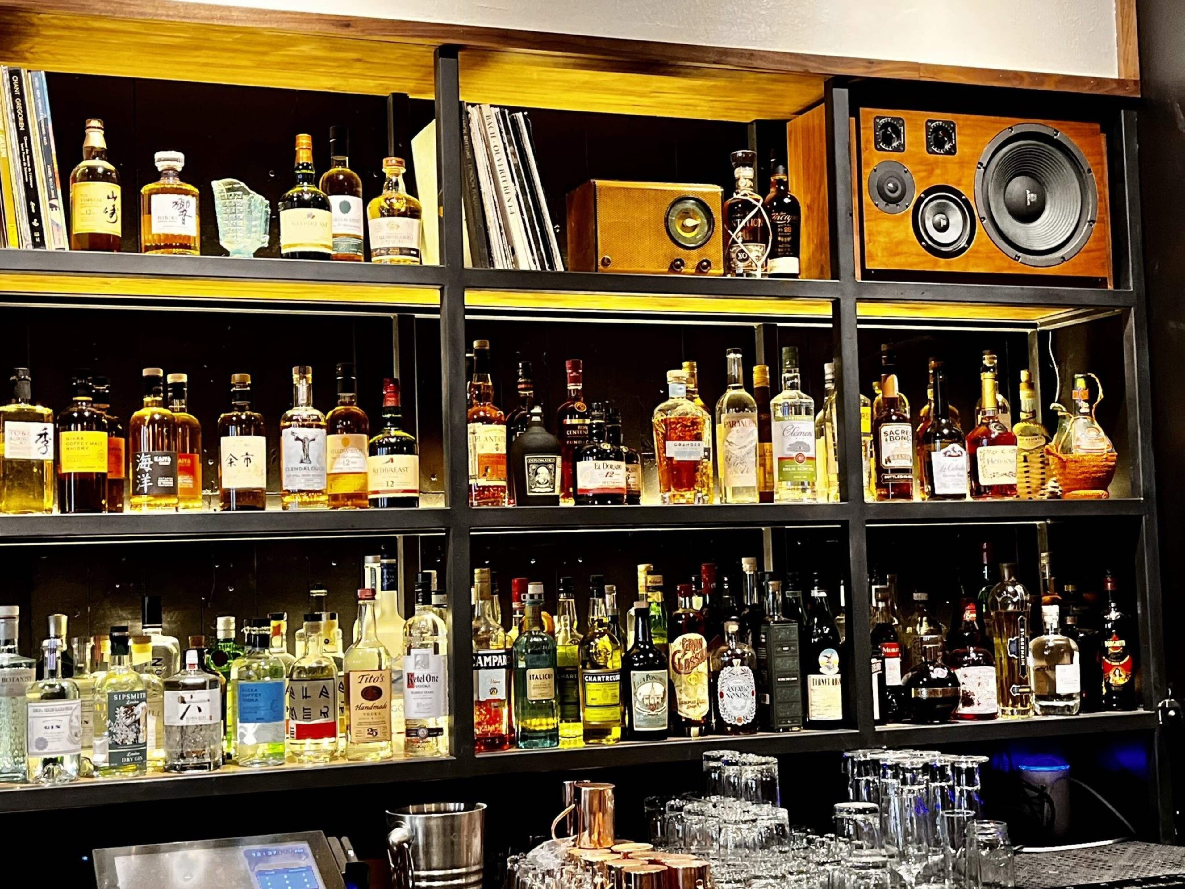 Shelves filled with various liquor bottles, vinyl records, a vintage radio, a large wooden speaker, and a bar counter with glasses and bar tools.